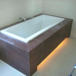RemontLux - Wanna obudowana ciemnobrązowymi płytkami, z podświetleniem LED u dołu, widoczna drabina z fragmentem spodni w rogu
