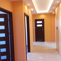 RemontLux - Korytarz po remoncie z pomarańczowymi ścianami, trzema ciemnymi drzwiami z przeszkleniami, podwieszanym sufitem z oświetleniem punktowym i jasnymi płytkami podłogowymi.