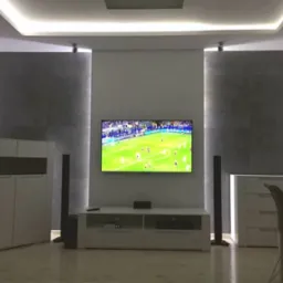 Nowoczesny salon z szarymi panelami ściennymi, podświetlanym sufitem i telewizorem wyświetlającym mecz piłkarski, minimalistyczne meble w kolorze białym i czarnym.