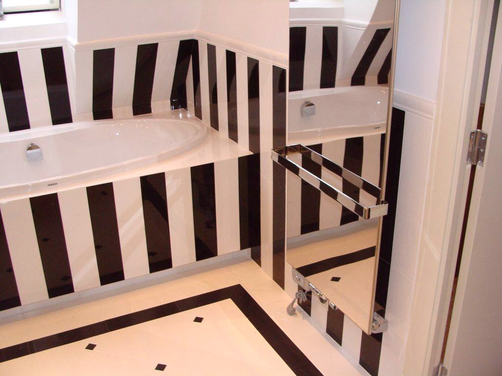 Nowoczesna łazienka z wanną obudowaną w czarno-białe pasy, pasy kontynuowane na ścianach, podłoga w jasnych płytkach z czarnymi akcentami, lustro z wieszakiem na ręczniki.
