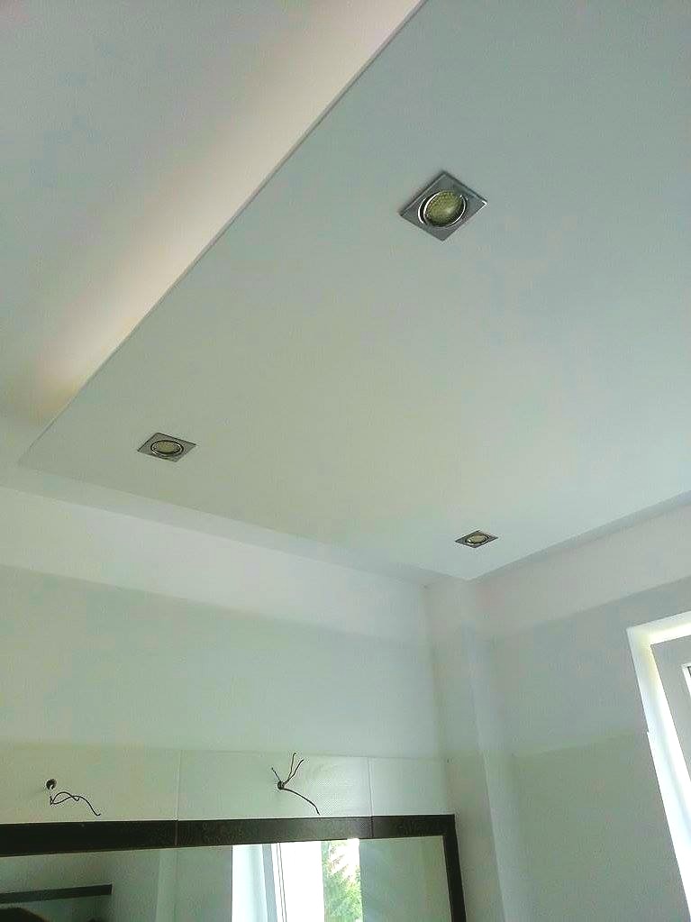 Biały sufit podwieszany z trzema kwadratowymi oprawami halogenowymi, widoczne również białe ściany i fragment lustra z czarną ramą w łazience po remoncie.