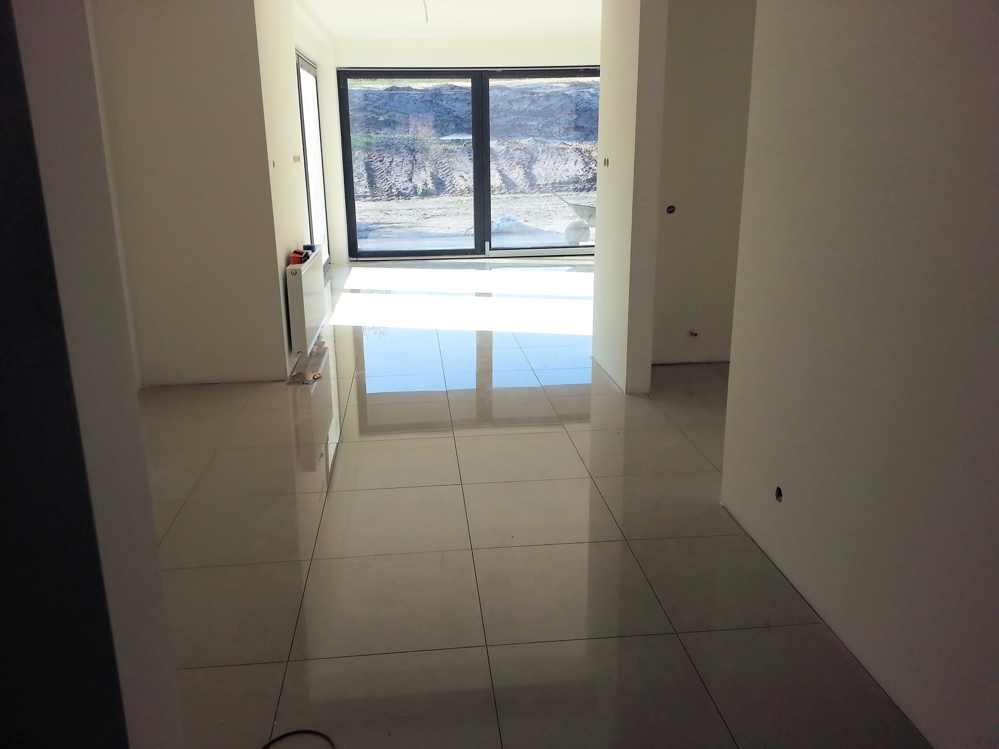 Wnętrze w stanie deweloperskim z jasnymi, błyszczącymi płytkami podłogowymi, świeżo pomalowanymi ścianami na biało i dużym oknem przesuwnym z widokiem na teren budowy.