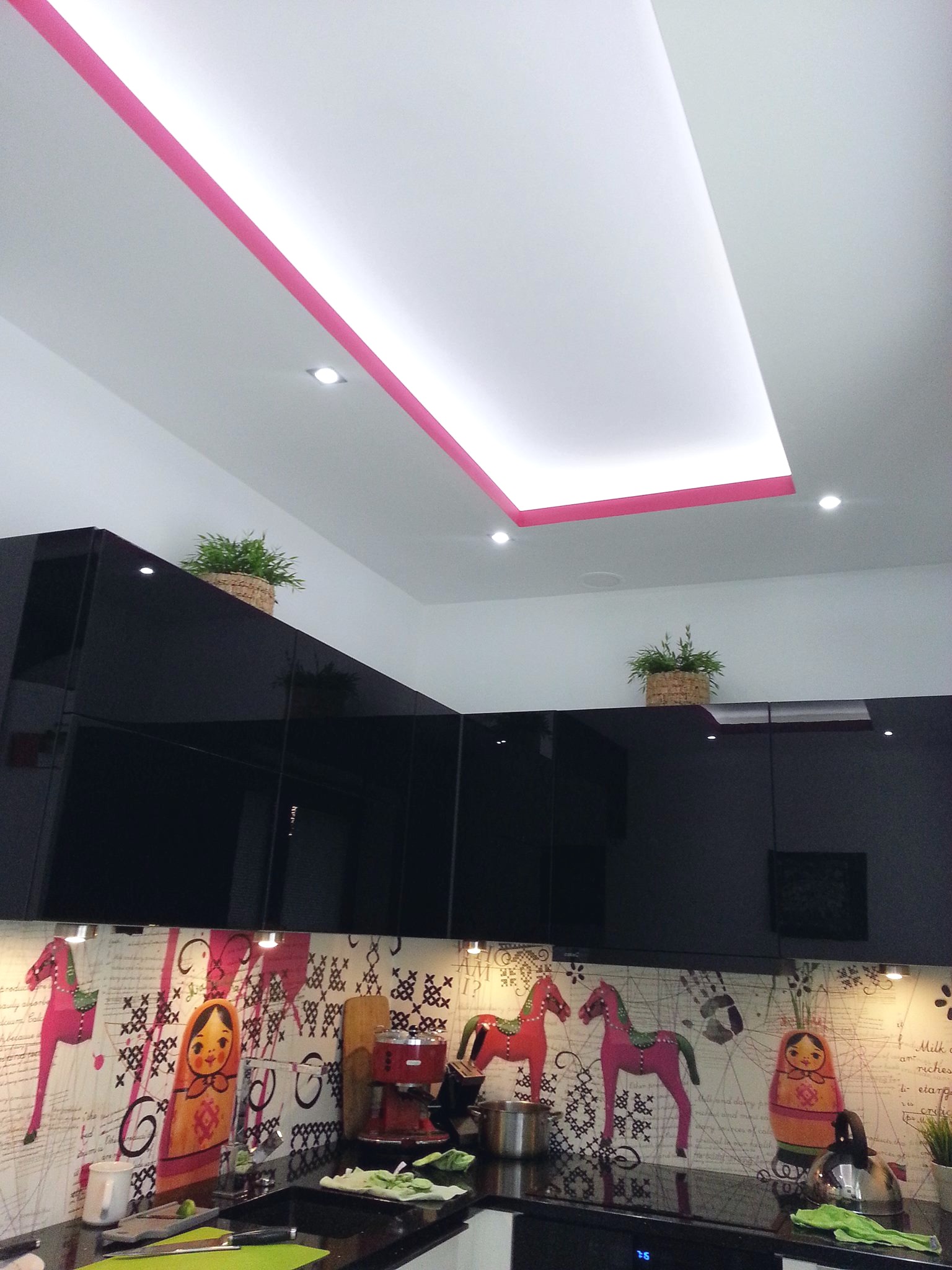 Nowoczesna kuchnia z czarnymi, błyszczącymi szafkami, dekoracyjnym panelem nad blatem z motywem rosyjskich lalek i koni, oraz podwieszanym sufitem z oświetleniem obrysowanym różową lamówką.