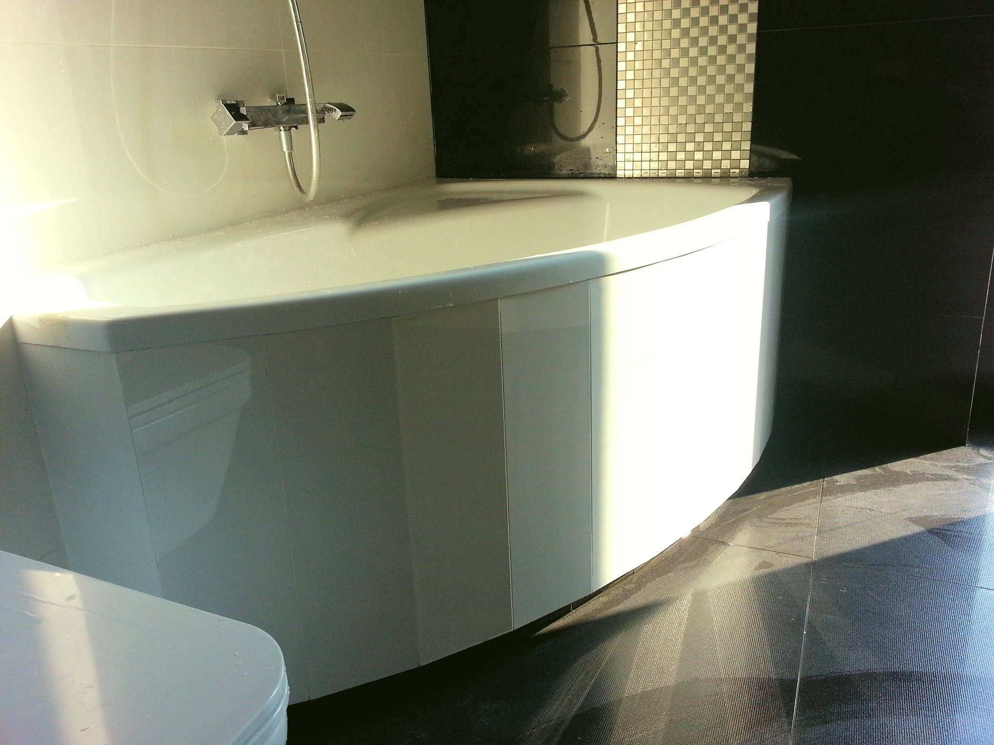 Nowoczesna, narożna wanna z hydromasażem, biała armatura, czarne płytki podłogowe i mozaika ścienna, minimalistyczny design, oświetlenie górne.