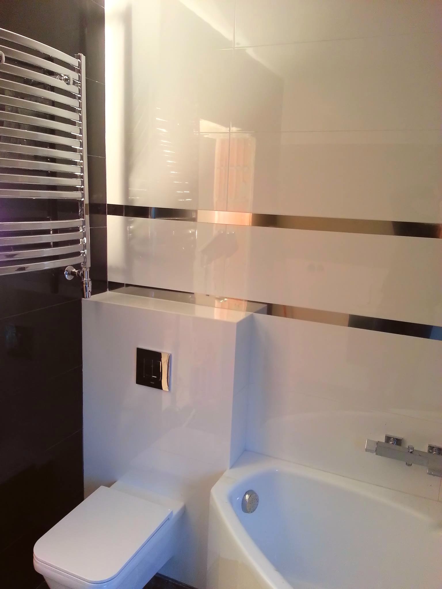 Nowoczesna łazienka w czarno-białej kolorystyce z połyskującymi płytkami, chromowanym grzejnikiem drabinkowym i minimalistyczną armaturą, prezentująca efektowny kontrast kolorów i materiałów.