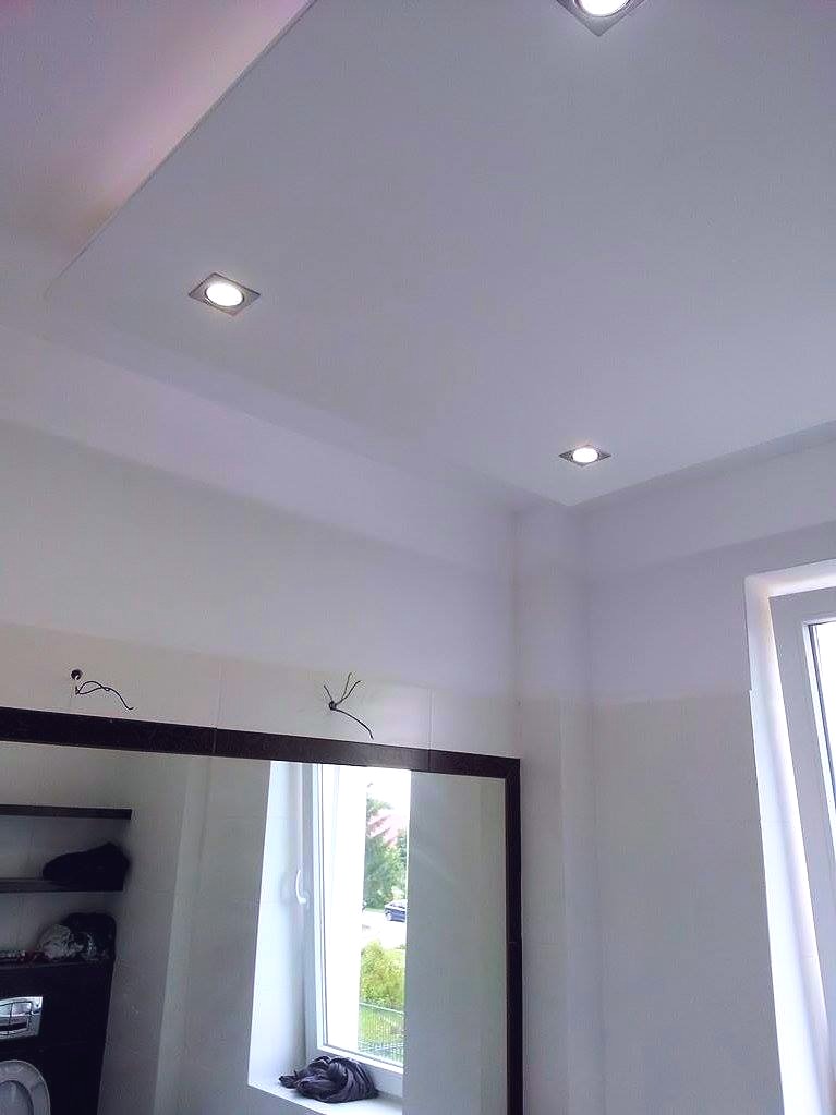 Biała łazienka z podwieszanym sufitem, oświetleniem punktowym i dużym lustrem z ciemną ramą, w którym odbija się okno.