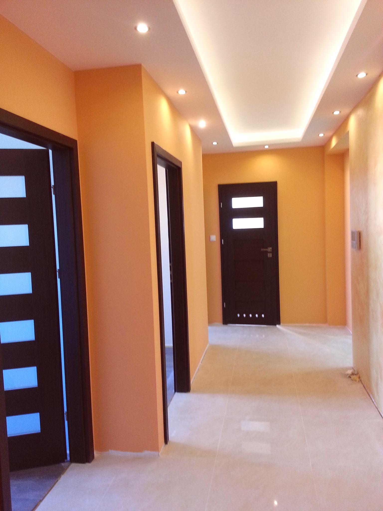 Korytarz po remoncie z pomarańczowymi ścianami, trzema ciemnymi drzwiami z przeszkleniami, podwieszanym sufitem z oświetleniem punktowym i jasnymi płytkami podłogowymi.