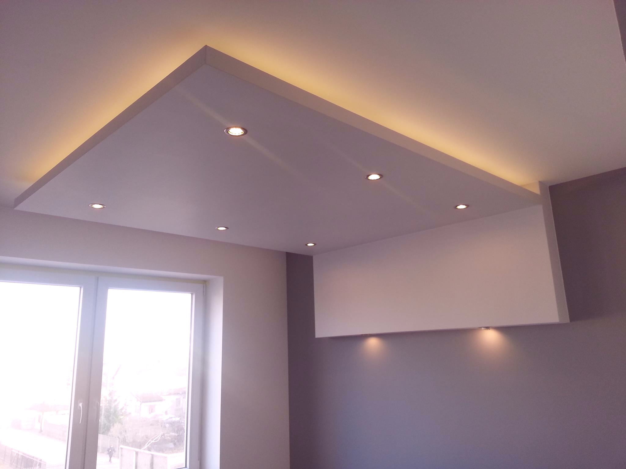 Nowoczesny sufit podwieszany z oświetleniem LED punktowym i taśmowym, widok z dołu na okno i szarą ścianę.