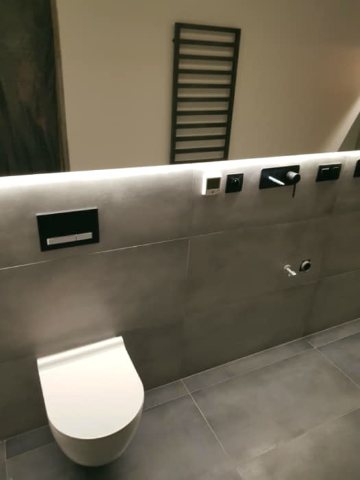 Nowoczesna łazienka wykończona w szarościach z podświetlanym lustrem, wiszącą toaletą i czarnym grzejnikiem drabinkowym.