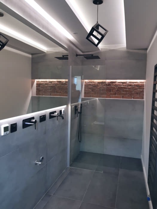 Nowoczesna łazienka z szarą płytką imitującą beton, oświetleniem LED w suficie, dekoracyjną ścianą z cegły i czarną armaturą oraz geometrycznymi lampami wiszącymi.