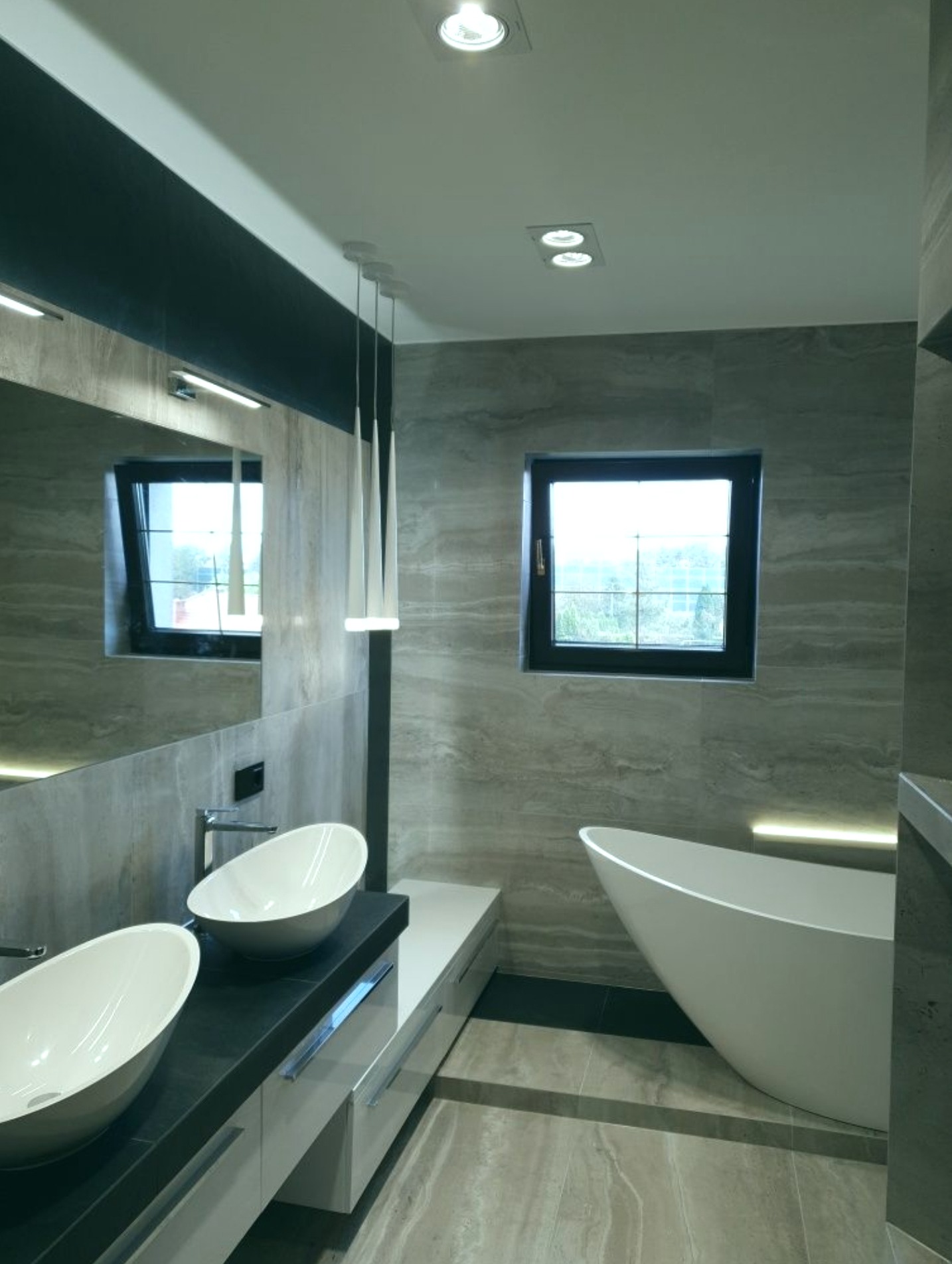 Nowoczesna łazienka z dwoma owalnymi umywalkami na czarnym blacie, wolnostojąca wanna przy oknie, szare płytki imitujące marmur na ścianach i podłodze, oświetlenie punktowe sufitowe i listwy LED.
