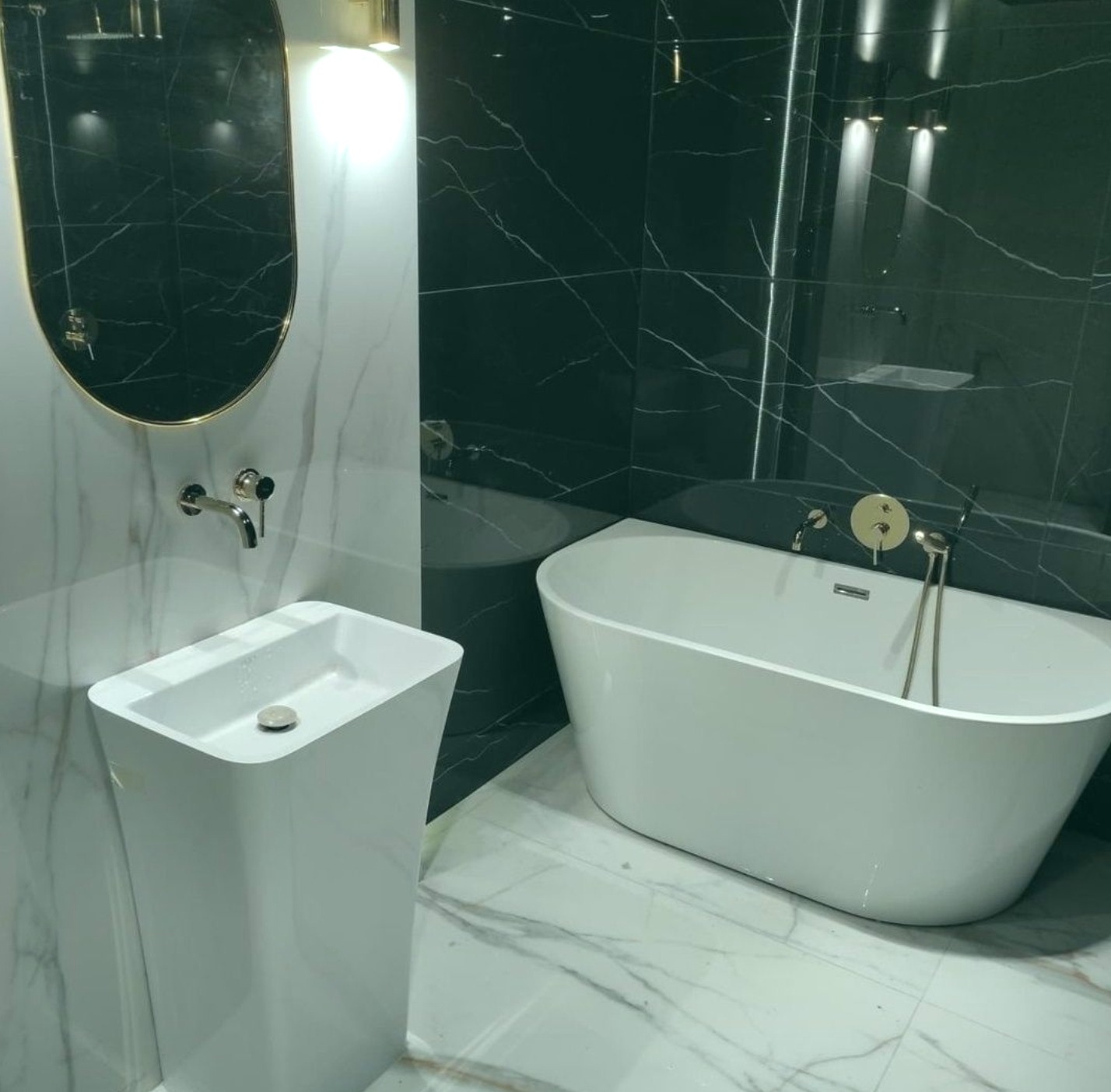 Nowoczesna łazienka z wolnostojącą wanną, umywalką na postumencie i czarno-białymi marmurowymi płytkami, oświetlona złotymi akcentami.