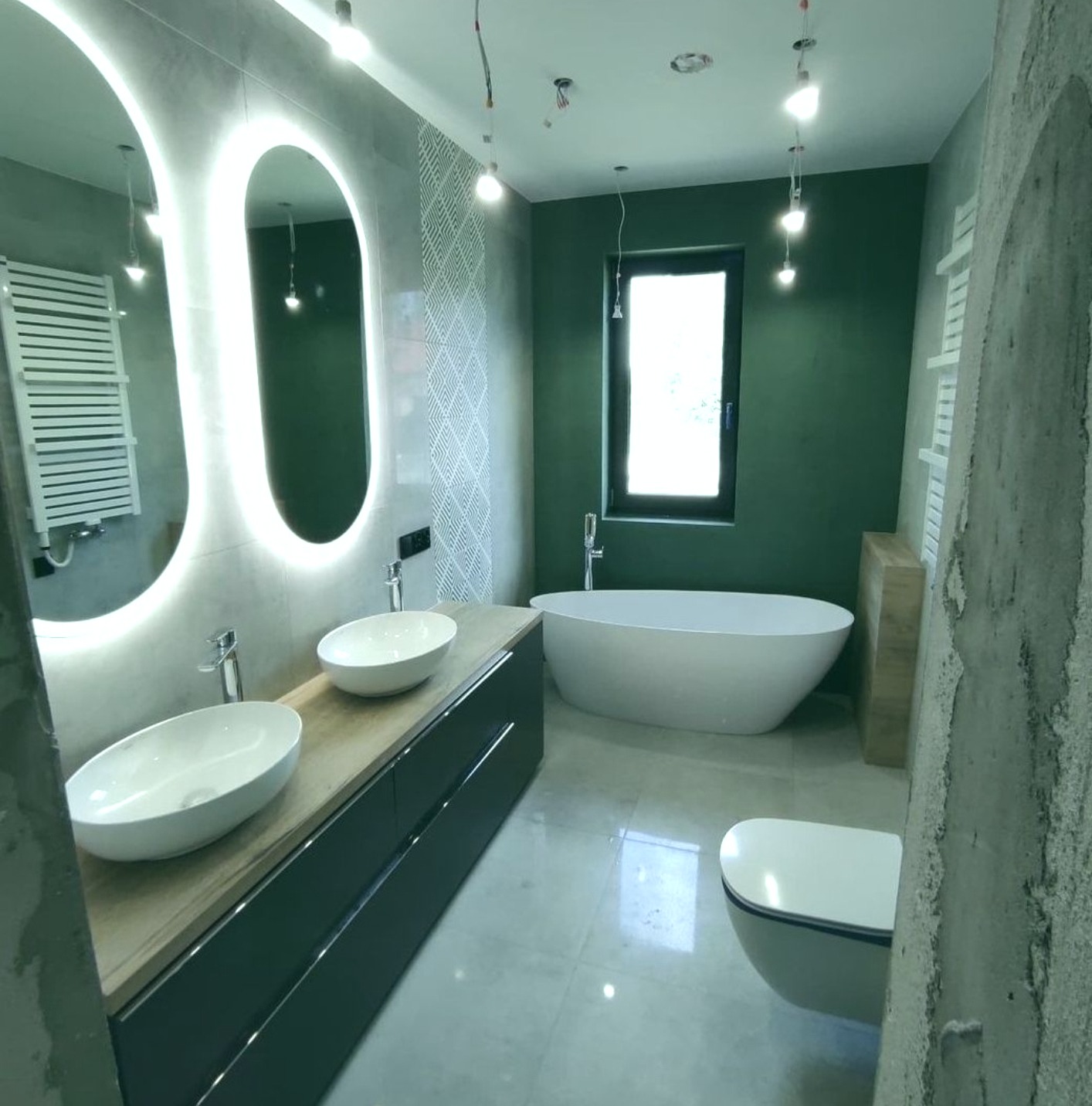 Nowoczesna łazienka z podwójną umywalką nablatową, owalnymi lustrami z podświetleniem LED, wolnostojącą wanną i zielonymi ścianami, w trakcie wykańczania.