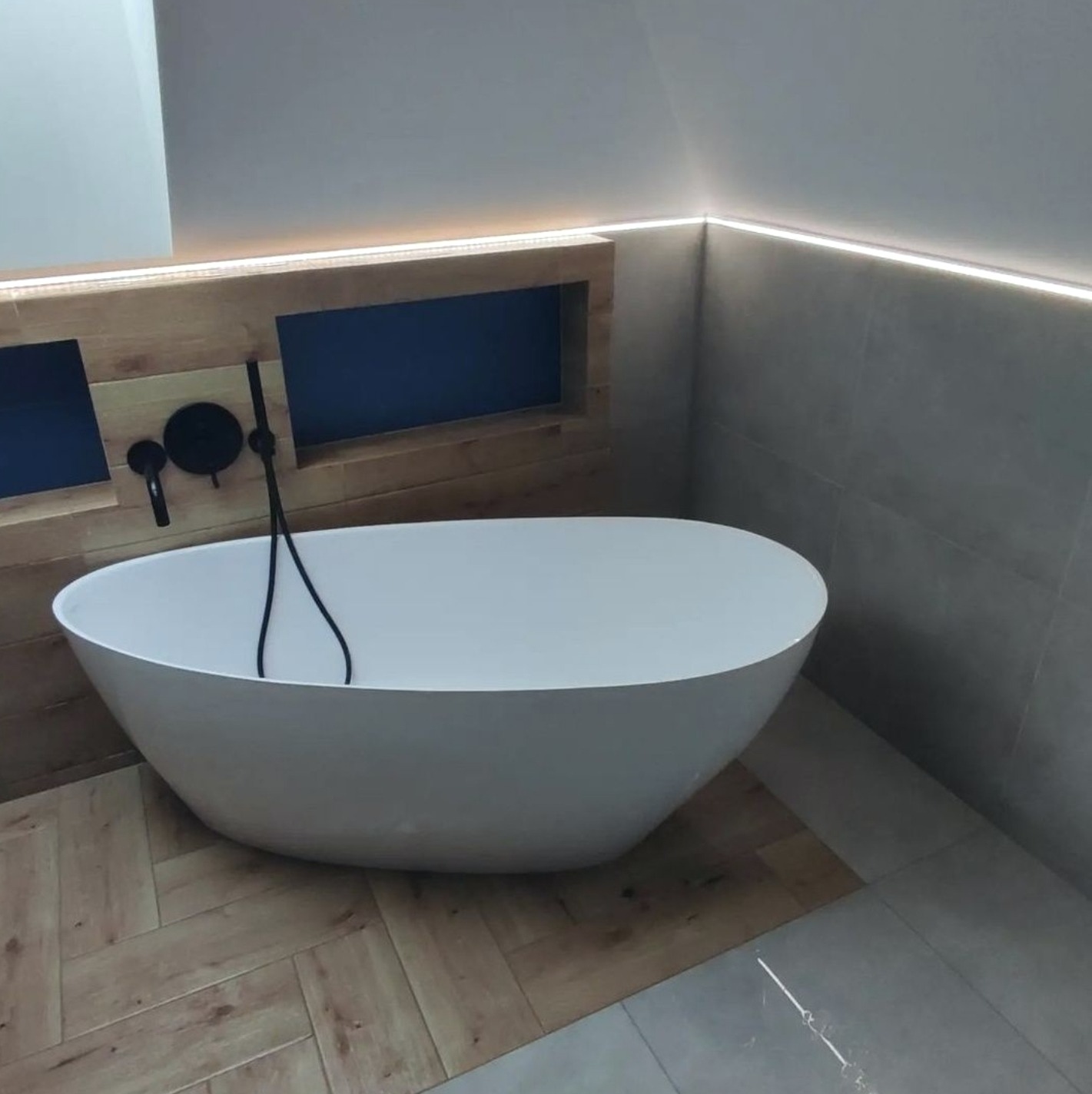 Wolnostojąca wanna w nowoczesnej łazience z drewnopodobną podłogą ułożoną w jodełkę i szarą ścianą z podświetleniem LED.