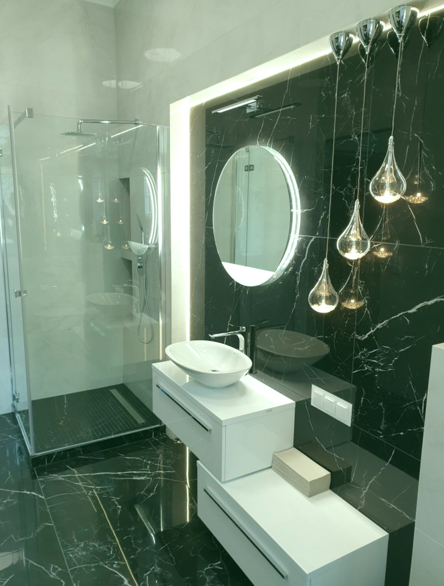 Nowoczesna łazienka z czarnymi marmurowymi płytkami, podświetlanym okrągłym lustrem, wiszącą armaturą, szklaną kabiną prysznicową i dekoracyjnymi lampami w kształcie kropli.