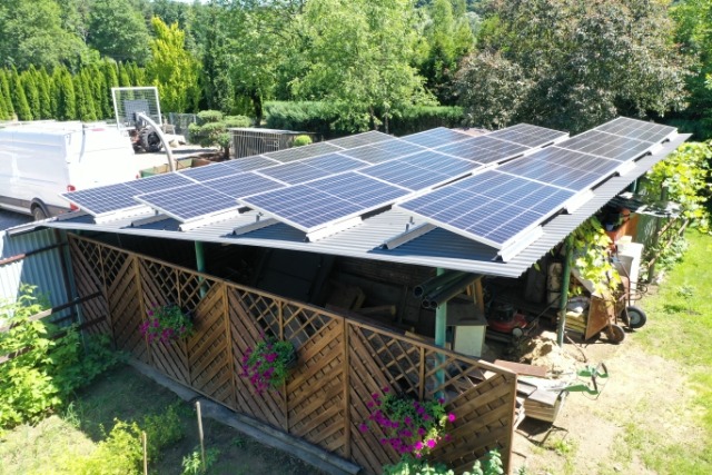 Instalacja paneli słonecznych na dachu wiaty z drewnianym ogrodzeniem i kwiatami, na tle zieleni.