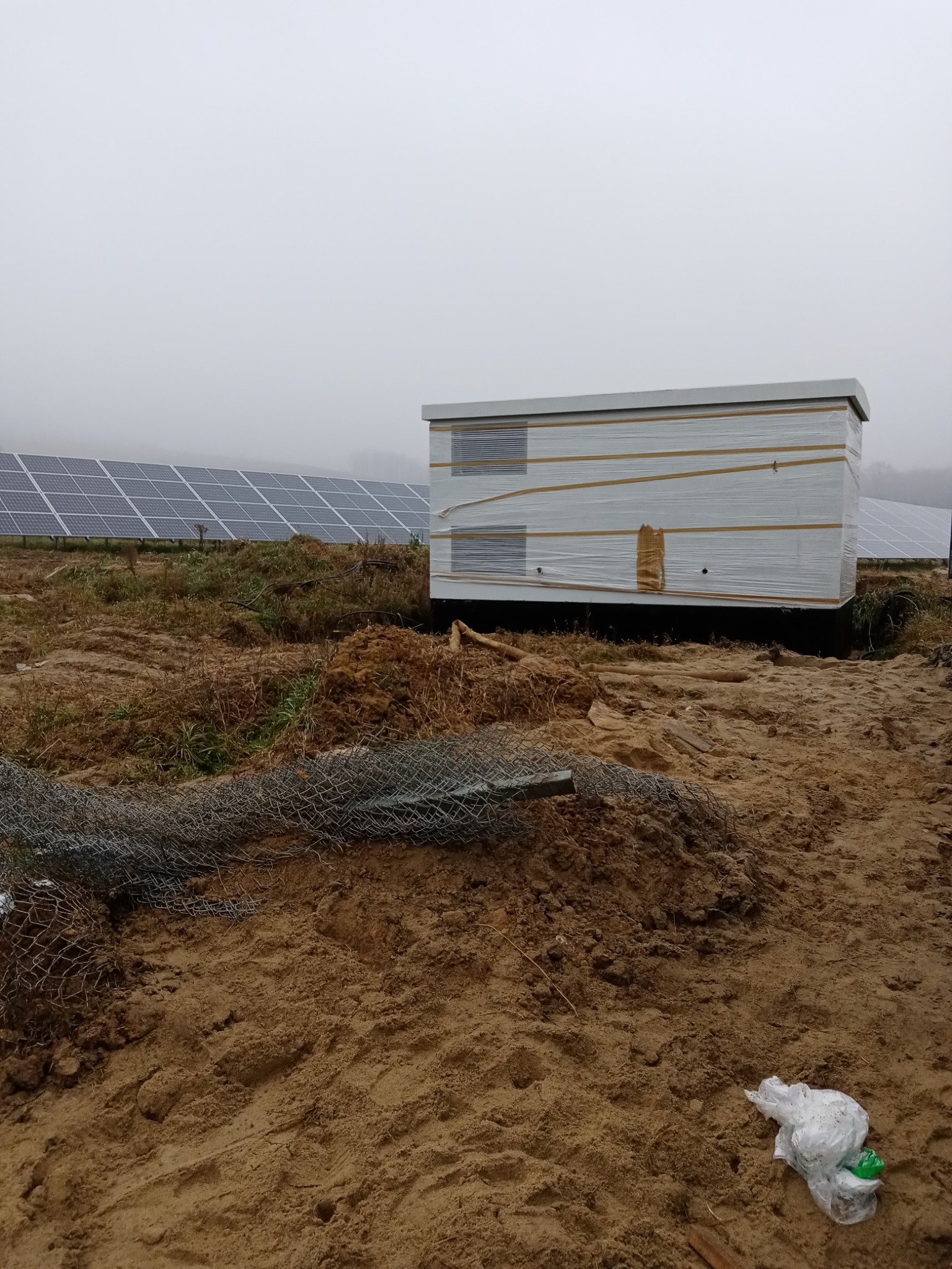 Nowy, owinięty folią magazyn energii na placu budowy, z panelami słonecznymi w tle, w pochmurny dzień.