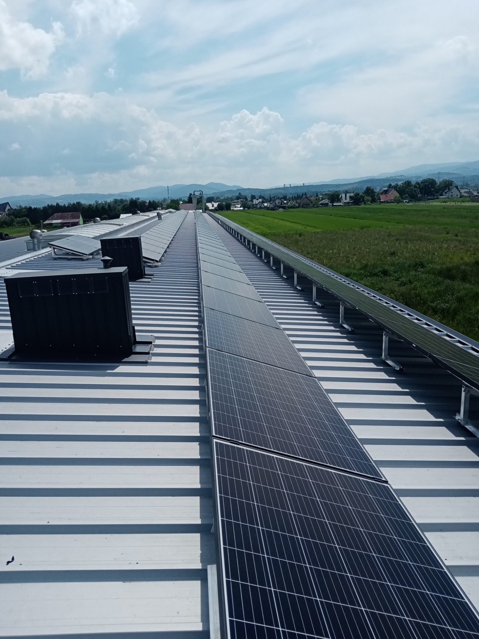 Instalacja paneli słonecznych na dachu przemysłowym z widokiem na zielone pola i zabudowania w oddali pod błękitnym niebem z chmurami.