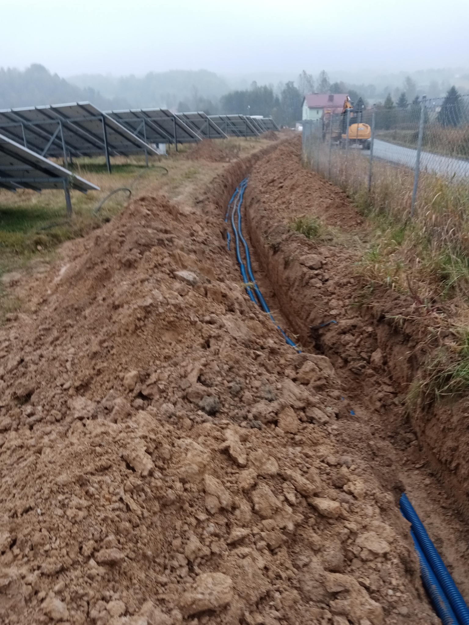 Wykop w ziemi z niebieskimi rurami kablowymi, w tle panele słoneczne i koparka przy ogrodzeniu.