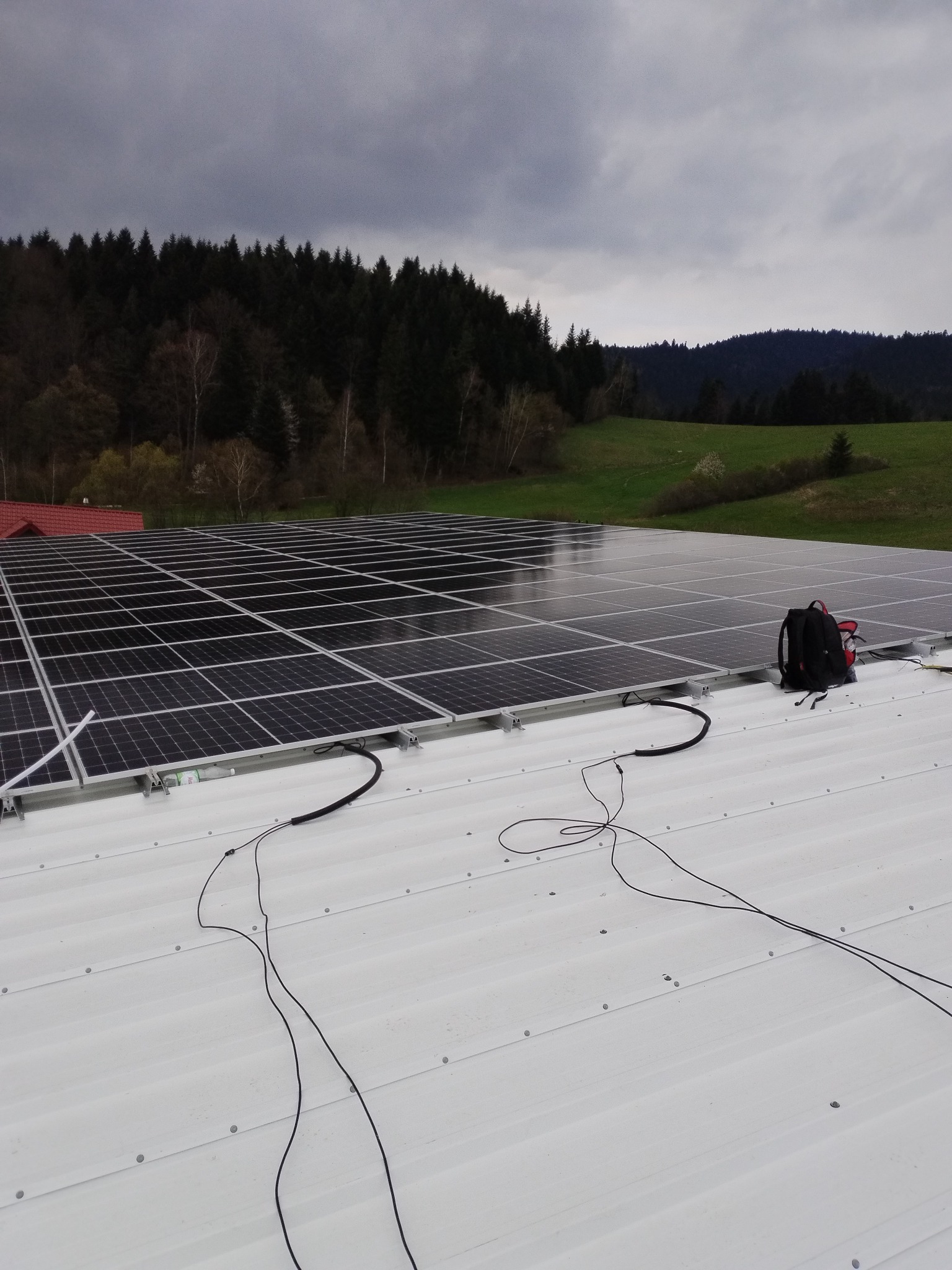 Instalacja paneli słonecznych na dachu z widocznymi kablami i plecakiem, w tle zalesione wzgórza pod pochmurnym niebem.