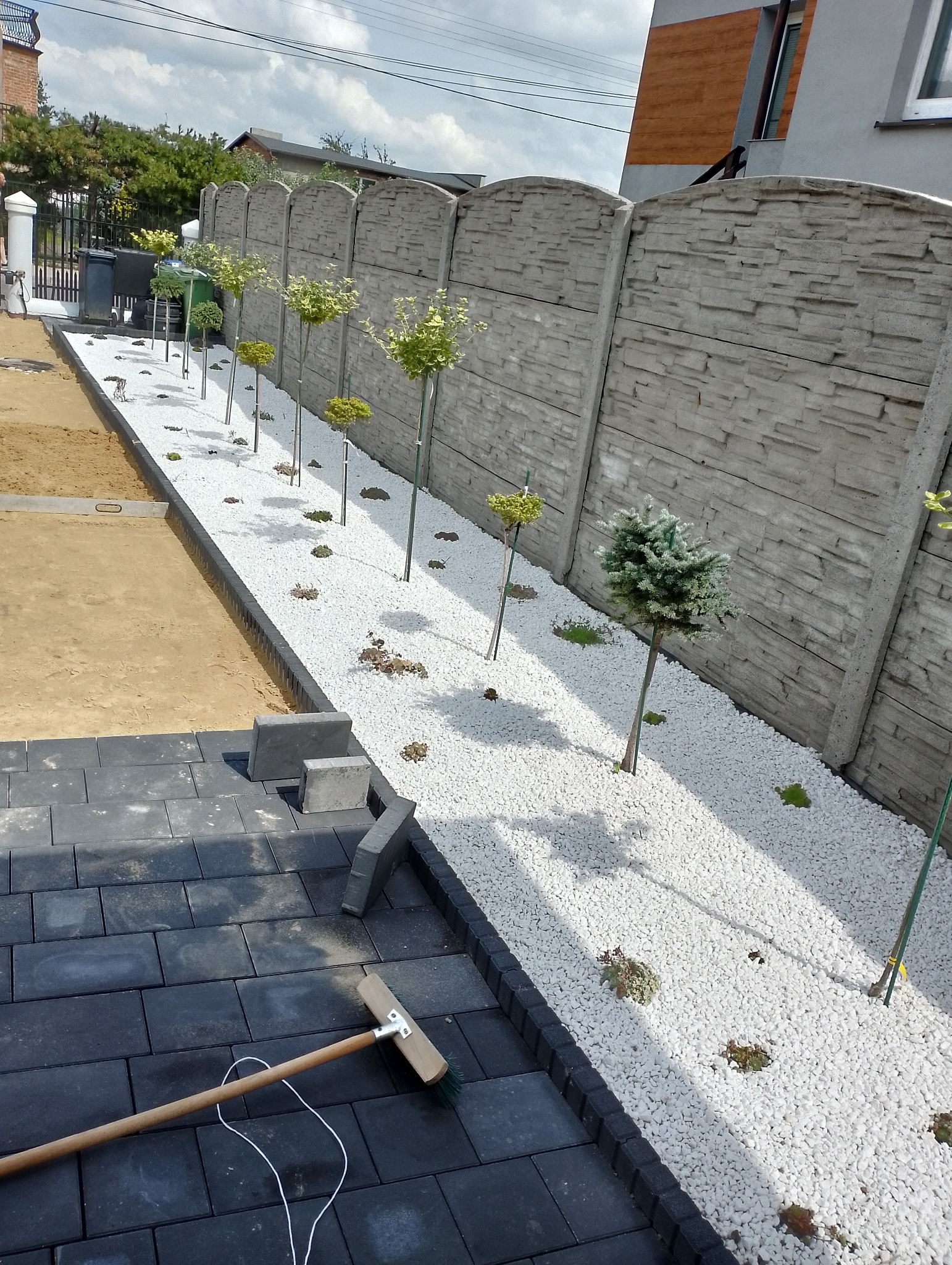 Fragment ogrodu w trakcie realizacji: ułożona część nawierzchni z ciemnej kostki brukowej, obok pas białego kamienia z nasadzeniami młodych drzewek i krzewów, widoczny betonowy płot i narzędzie...