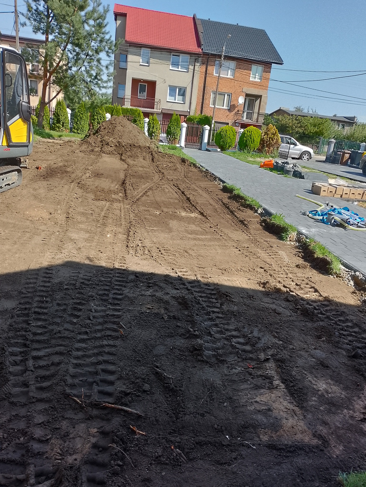 Przygotowanie terenu pod budowę chodnika z kostki brukowej. Widoczne ślady opon, hałda ziemi, częściowo ułożona kostka i budynki mieszkalne w tle.