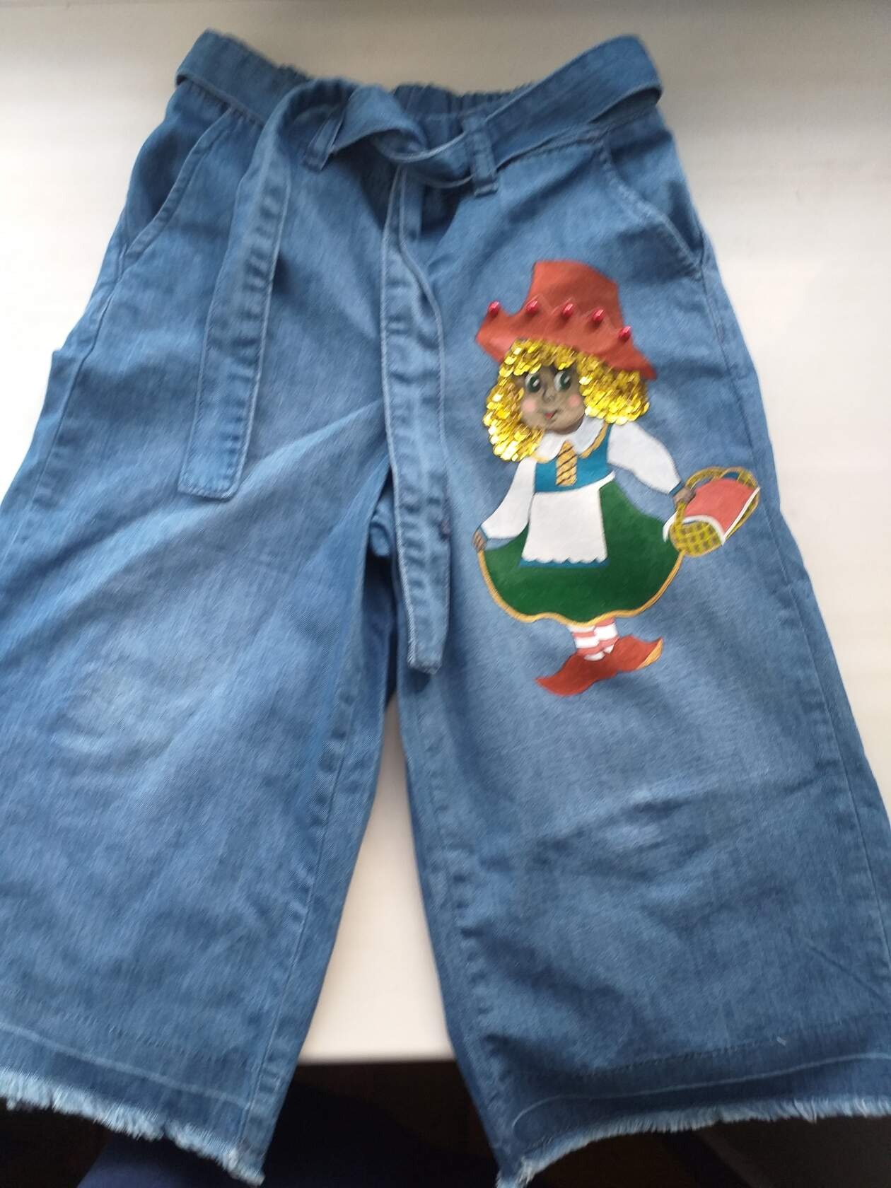 Dżinsowe spodnie-kuloty z ręcznie malowaną postacią dziewczynki w kapeluszu i z koszykiem, wykończone frędzlami.