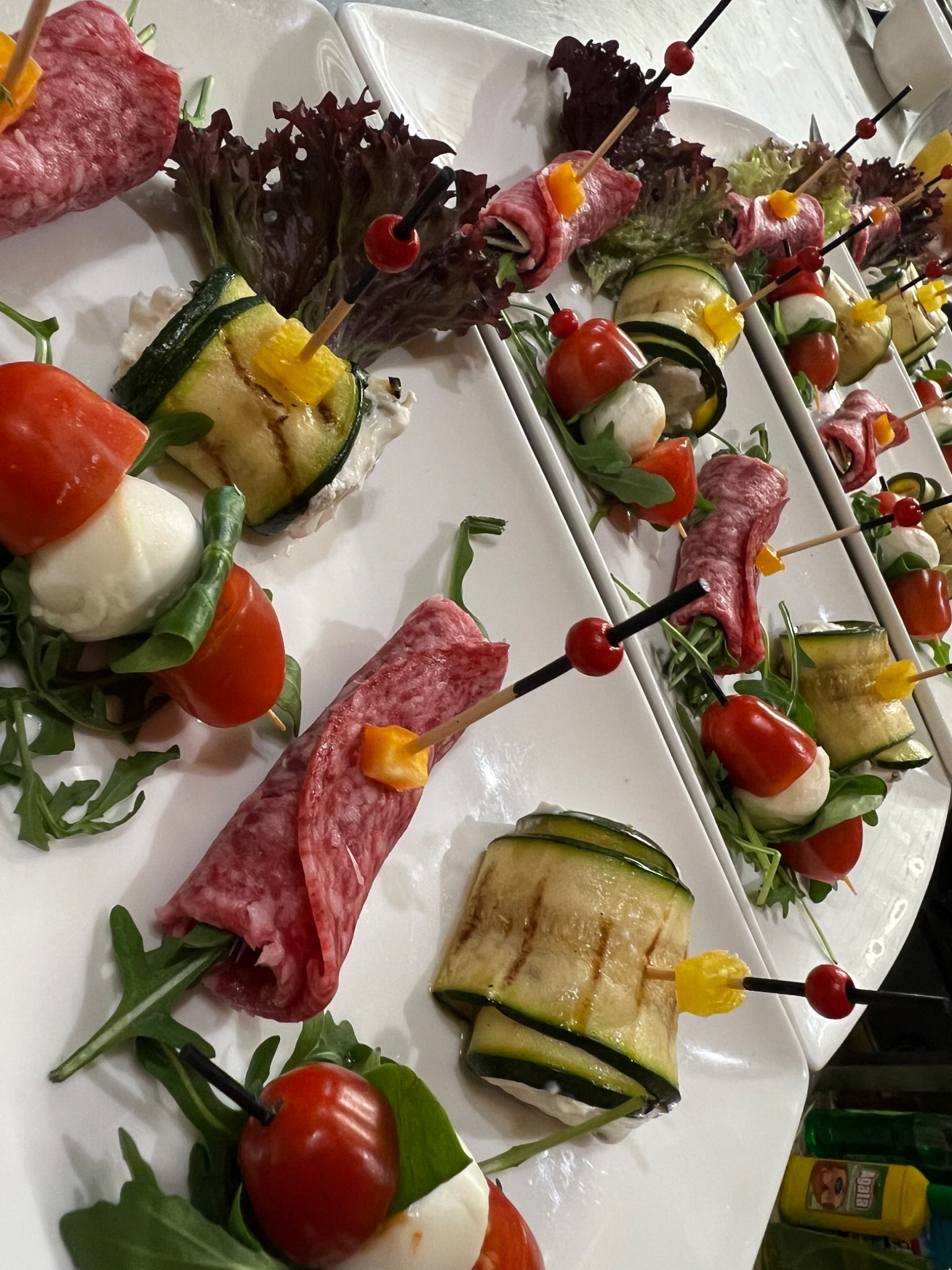 Rząd białych talerzy z apetycznymi przekąskami: salami w rulonie z rukolą, grillowana cukinia z kremowym serem, pomidorki cherry z mozzarellą, wszystko spięte ozdobnymi wykałaczkami.