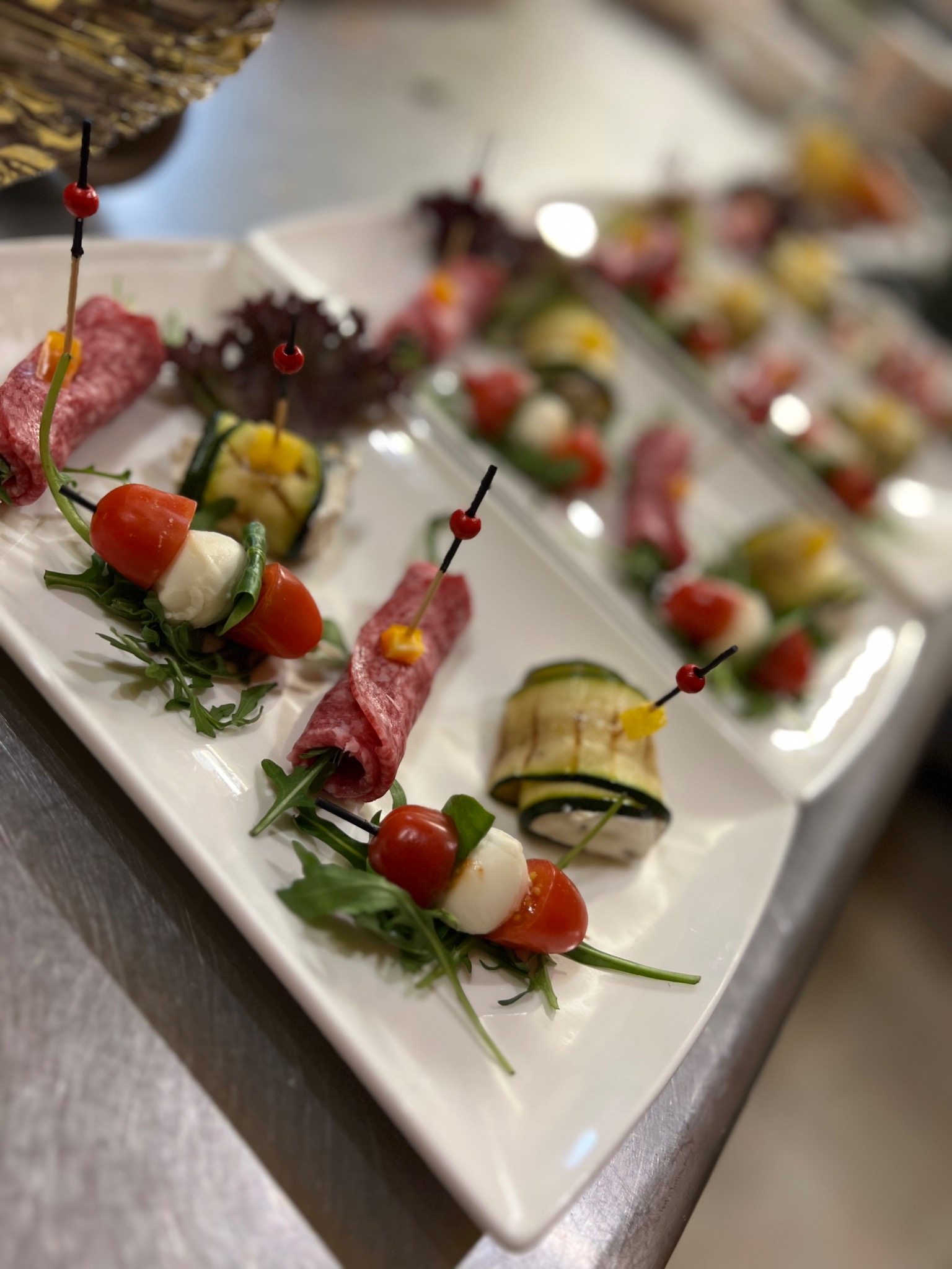 Elegancko podane przekąski na białych talerzach: salami w roladkach, koreczki caprese i grillowana cukinia z żółtą papryką, udekorowane gałązkami rukoli i czerwonymi ozdobnymi koralikami...