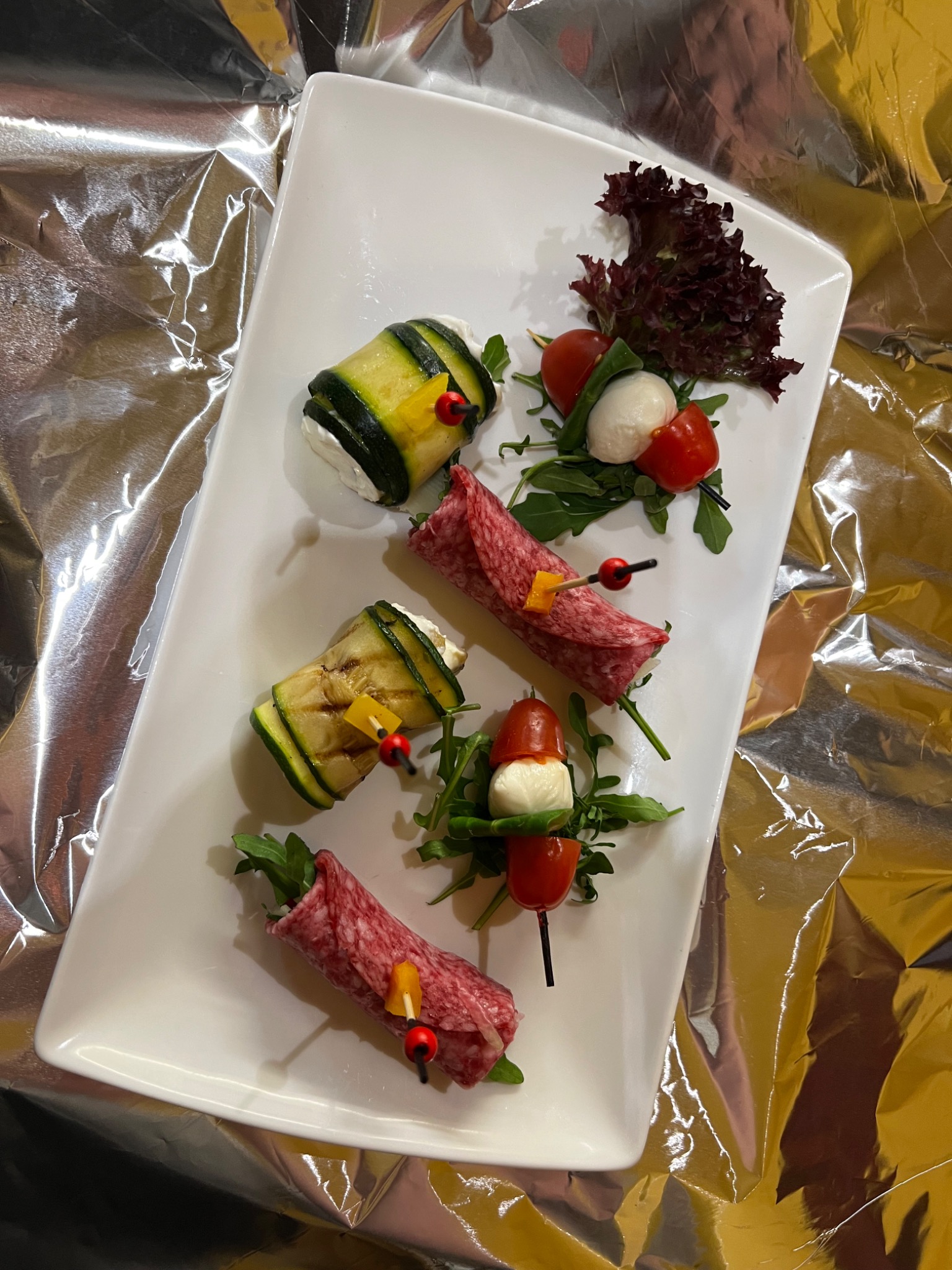 Elegancko podane przekąski na białym prostokątnym talerzu: roladki z grillowanej cukinii z serem, salami z dodatkami, koreczki caprese na tle błyszczącej folii.