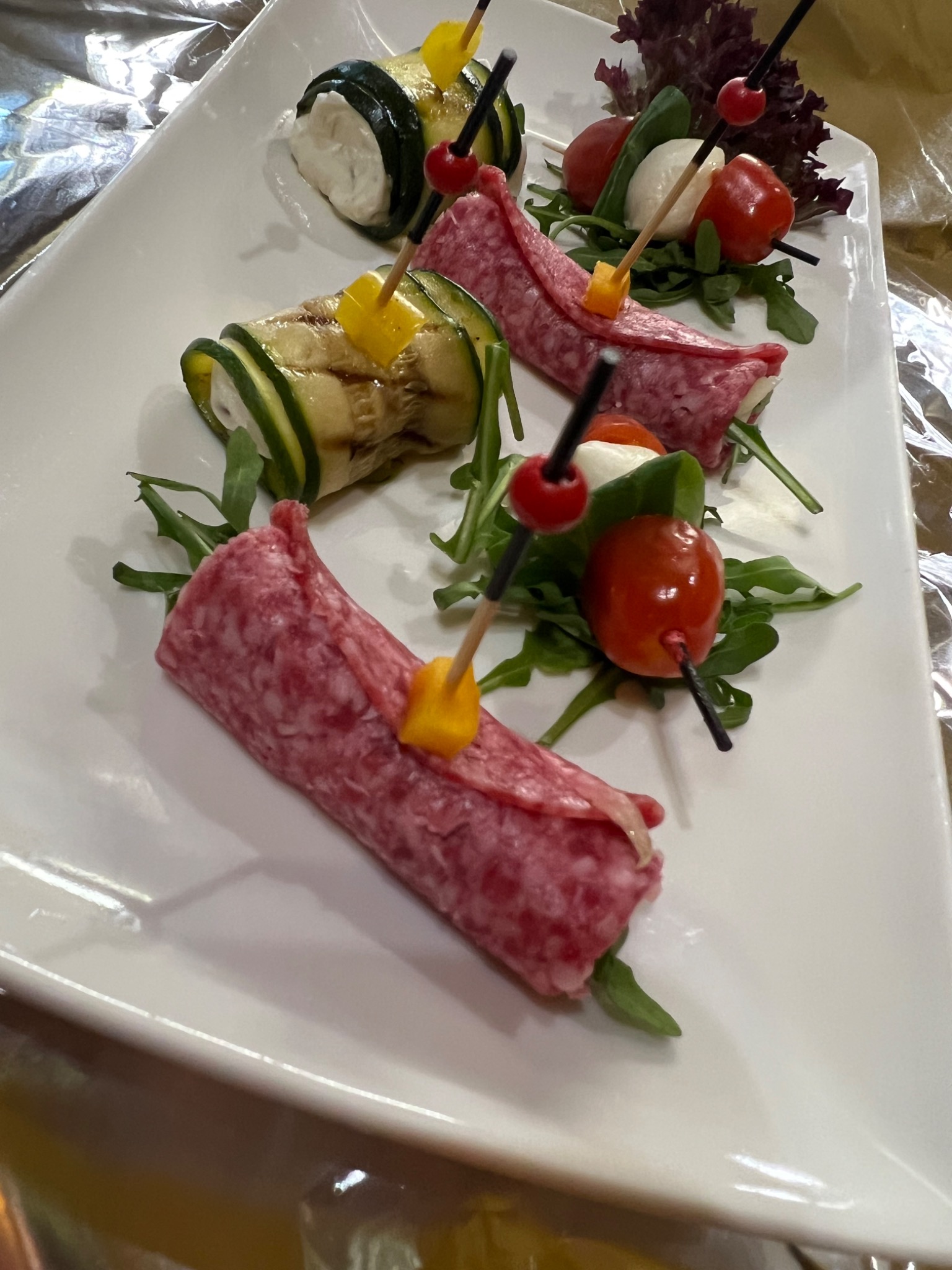 Elegancko podane przekąski na białym talerzu: roladki z salami i cukinii z dodatkiem świeżych ziół, pomidorków cherry i mini kulek mozzarelli, udekorowane kolorowymi szpikulcami.
