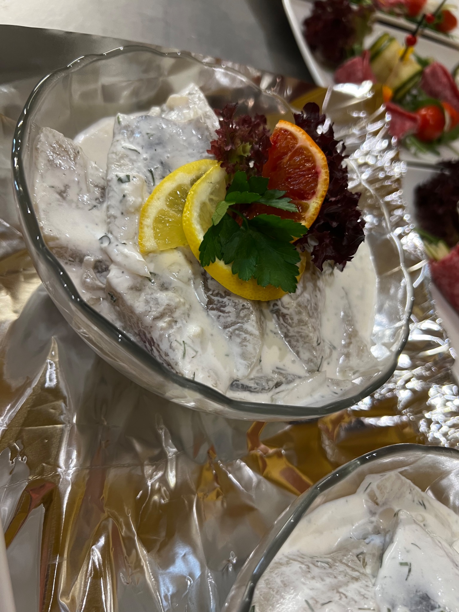 Śledzie w śmietanie udekorowane cytryną, krwistą pomarańczą i natką pietruszki, podane w szklanej miseczce na srebrnej folii.