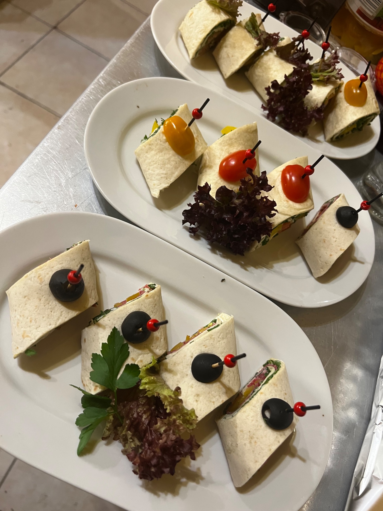 Trzy białe, owalne talerze z tortillami pokrojonymi w roladki, spięte czarnymi szpilkami z czerwonymi końcówkami, udekorowane sałatą i pomidorkami koktajlowymi w kolorach żółtym i czerwonym,...