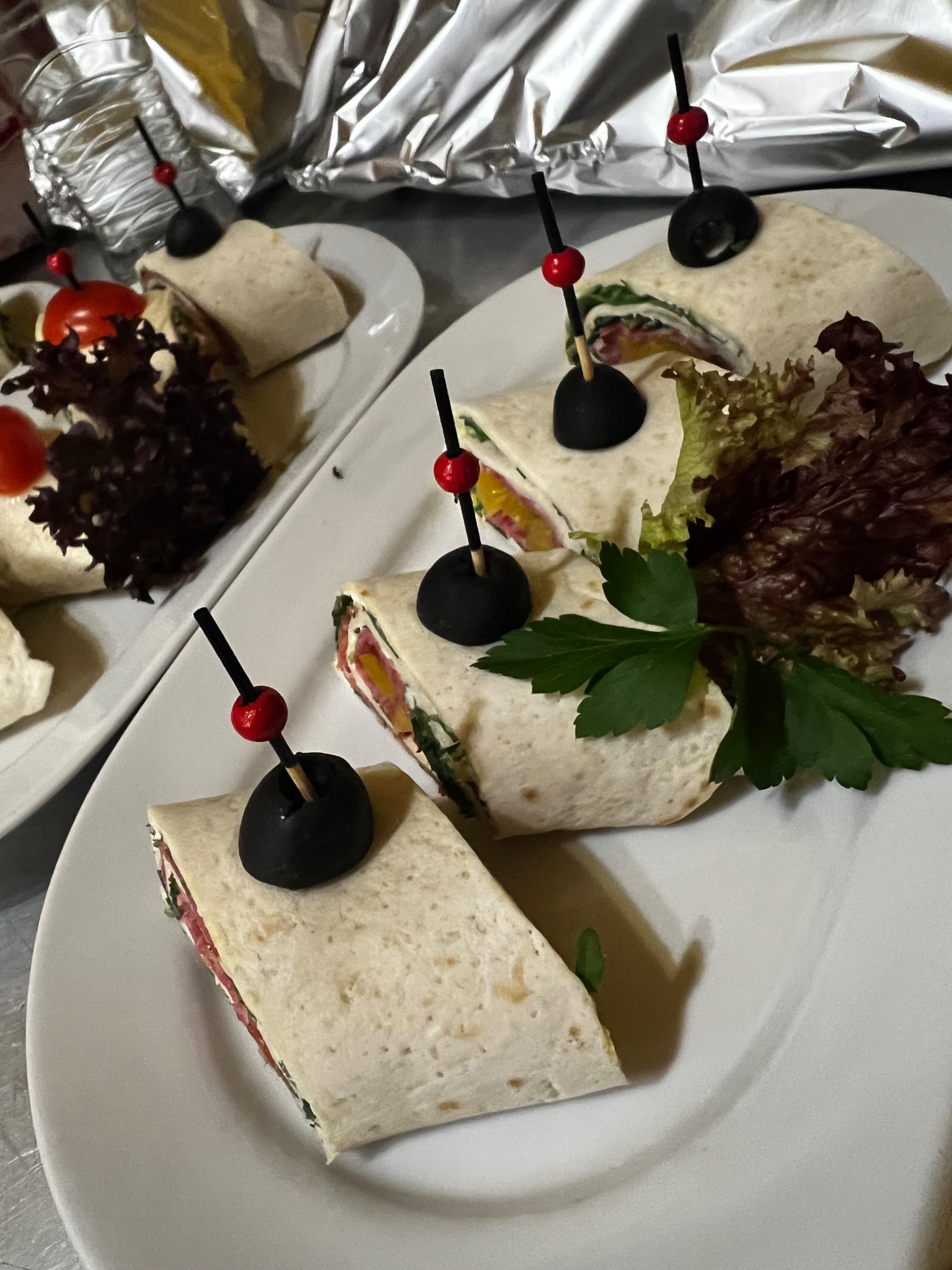 Tortilla wrap przekrojona na kawałki, udekorowana sałatą i oliwką na wykałaczce, ułożona na białym talerzu, w tle folia aluminiowa.