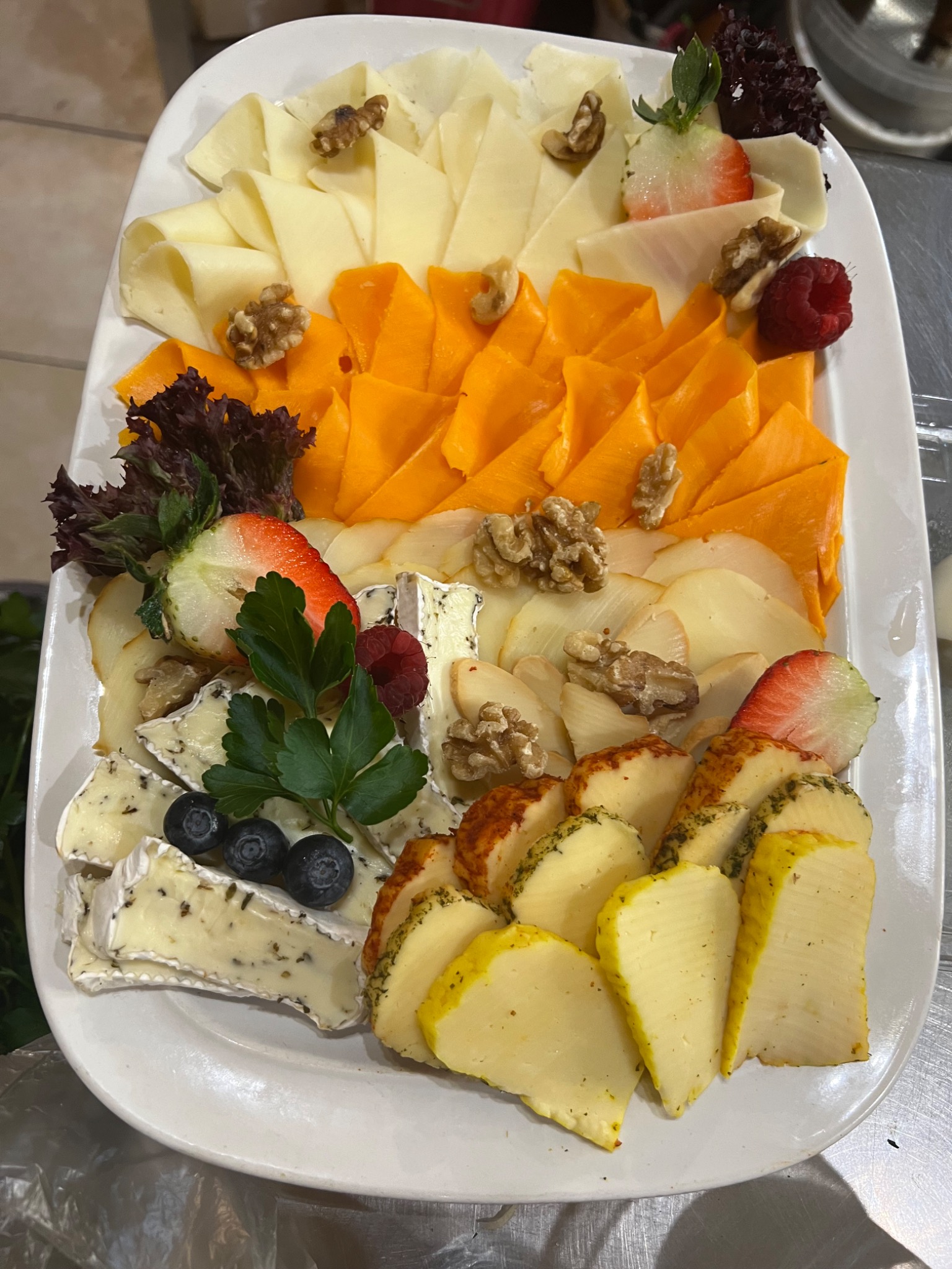 Elegancki talerz z różnorodnymi serami, orzechami włoskimi, malinami, borówkami, truskawkami i dekoracyjnymi liśćmi sałaty, ułożony na białym prostokątnym półmisku.