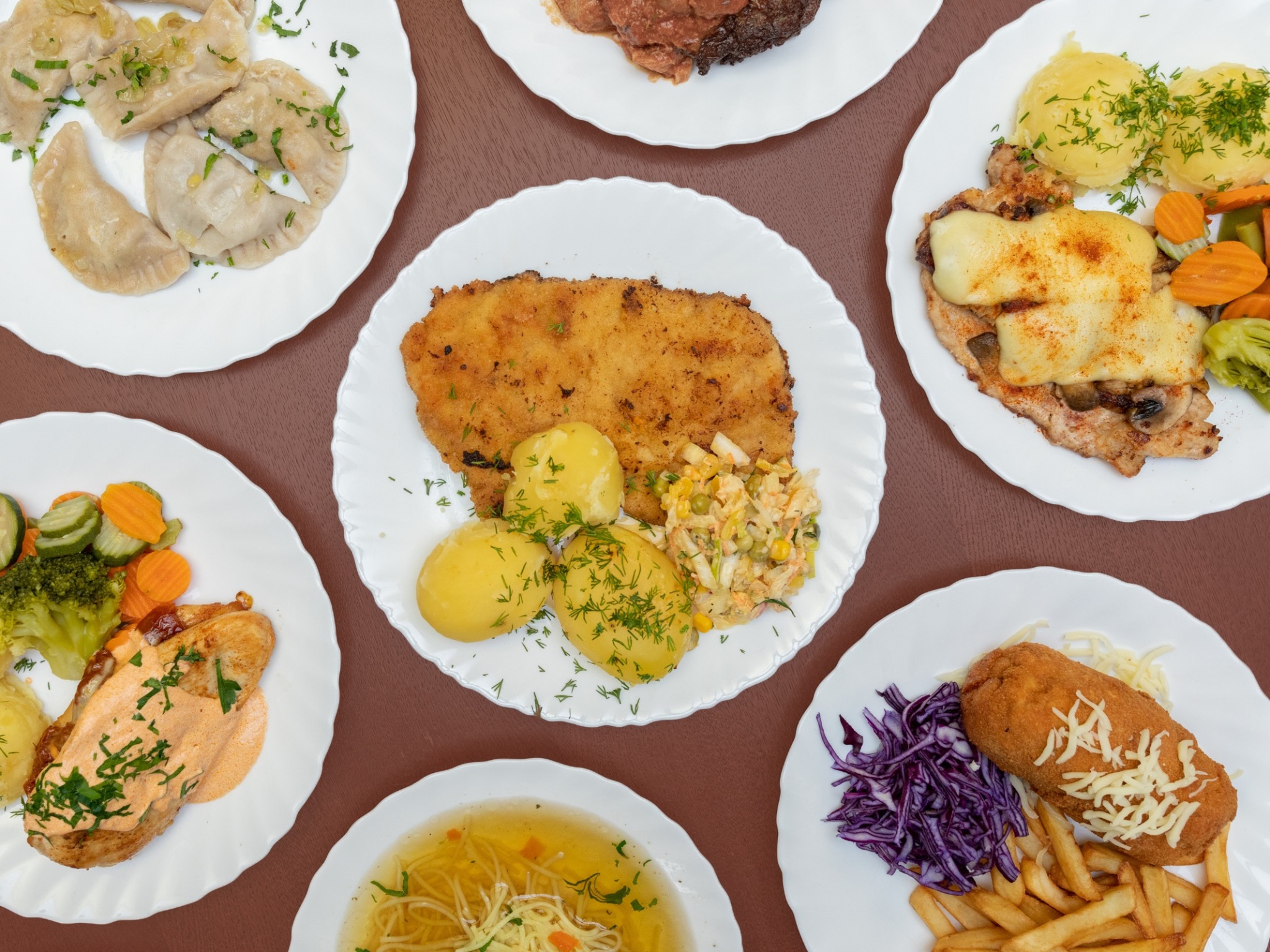 Różnorodne dania obiadowe na białych talerzach, w tym pierogi, kotlet schabowy z ziemniakami i surówką, devolay z frytkami i czerwoną kapustą, filet z kurczaka w sosie z warzywami, rosół...