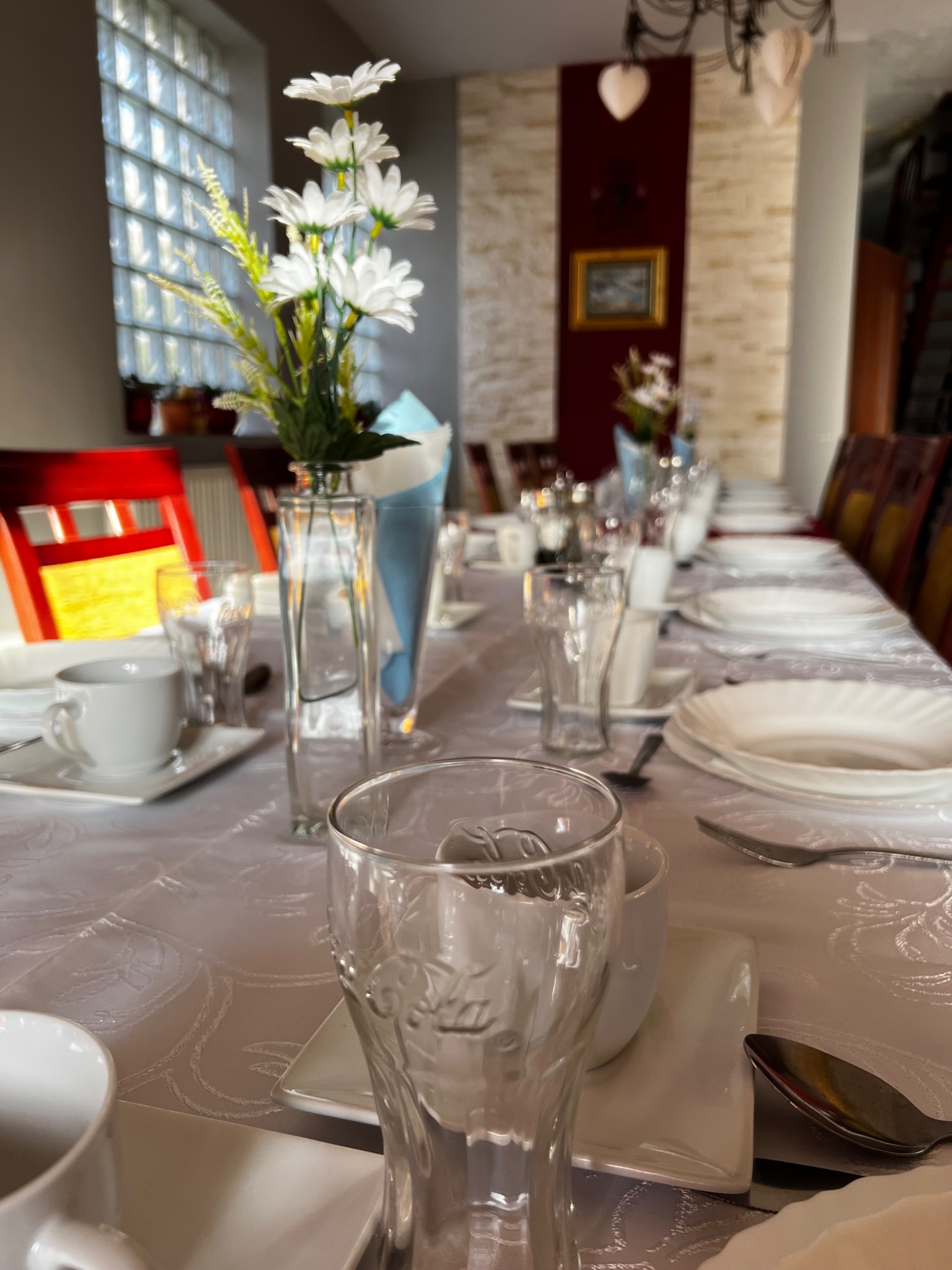 Ustawiony stół na uroczystość: biały obrus z wytłaczanym wzorem, biała zastawa, szklane wazony z białymi kwiatami, sztućce, szklanki z logo Coca-Cola, krzesła z czerwonymi oparciami w tle.