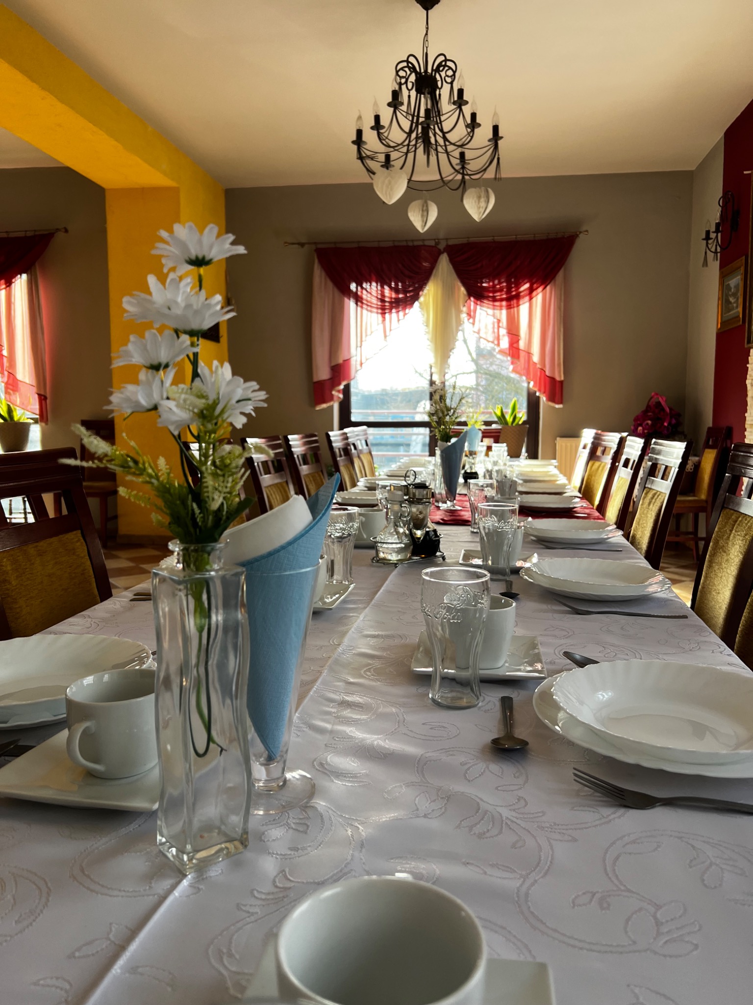 Długi stół nakryty białym obrusem z wytłaczanym wzorem, ustawiony dla wielu gości z białymi naczyniami, szklankami, sztućcami i dekoracjami z białych kwiatów w szklanych wazonach. W tle okna...