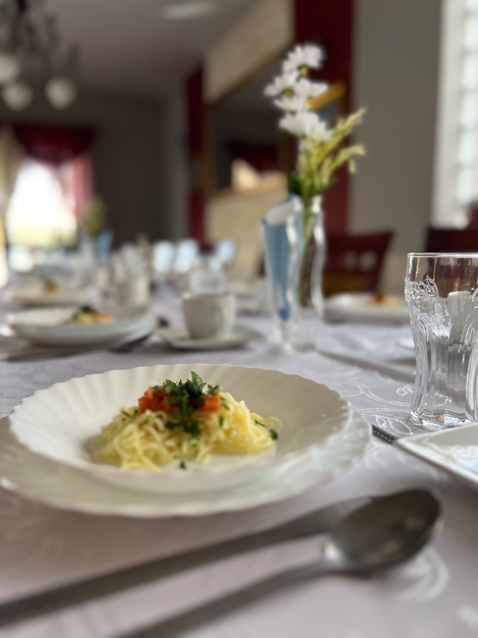 Uroczysty stół z nakryciem: talerz z makaronem, posypanym ziołami i pomidorami, szklanka, wazon z białymi kwiatami, biały obrus z delikatnym wzorem.