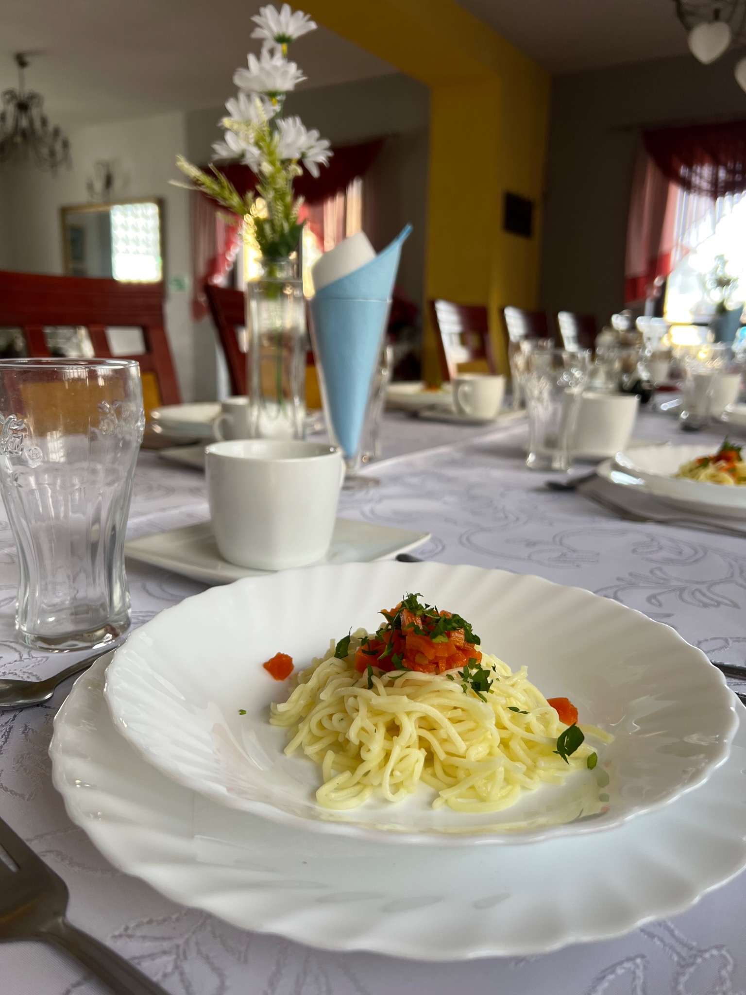Uroczysty stół nakryty białym obrusem, z talerzami z makaronem, filiżankami, szklankami i dekoracjami kwiatowymi, przygotowany na przyjęcie.