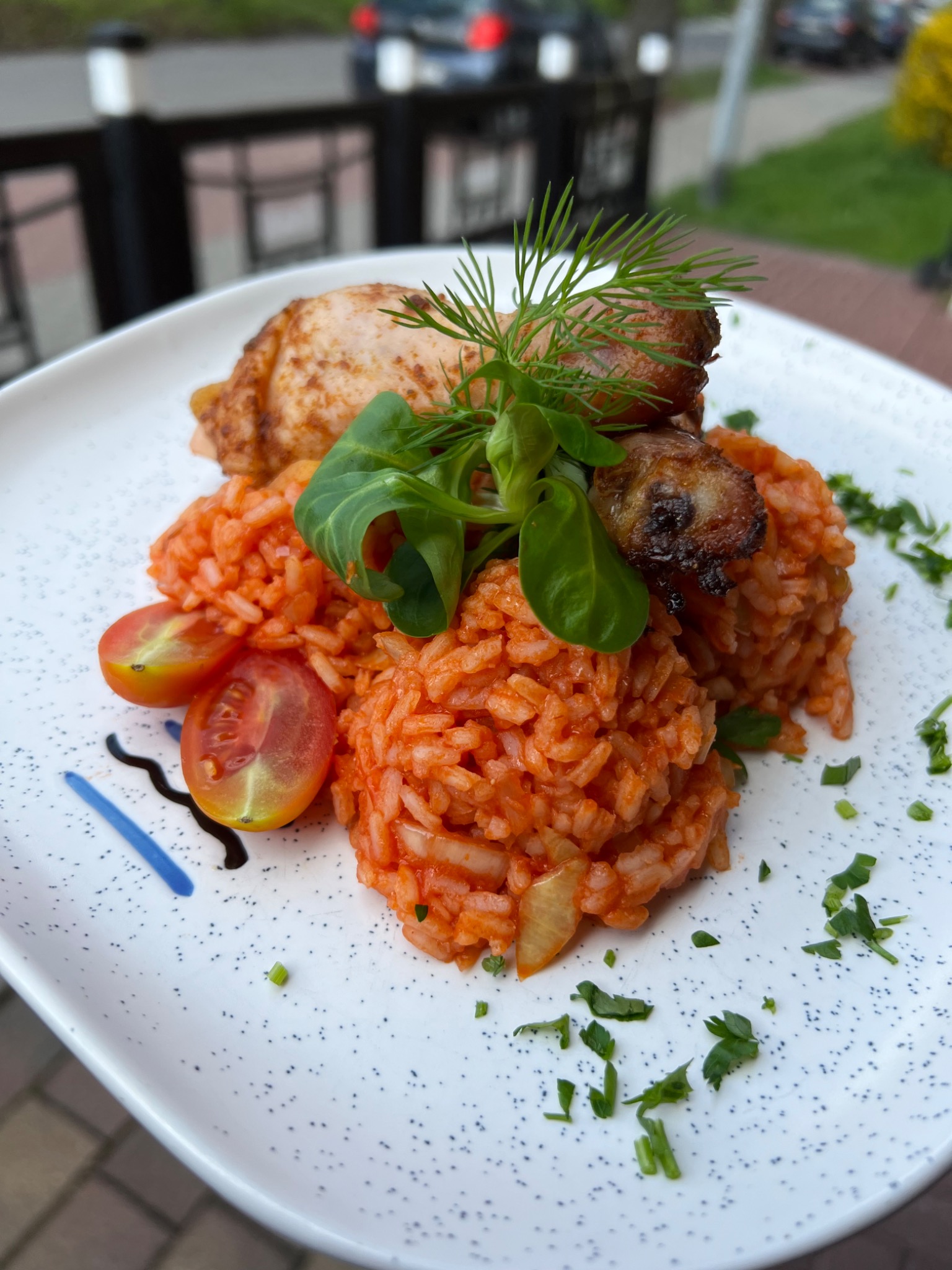 Elegancko podane danie: pieczone udo kurczaka, kulki ryżu w sosie pomidorowym, połówki pomidorków koktajlowych, dekorowane świeżymi ziołami i sosem balsamicznym na nakrapianym talerzu.