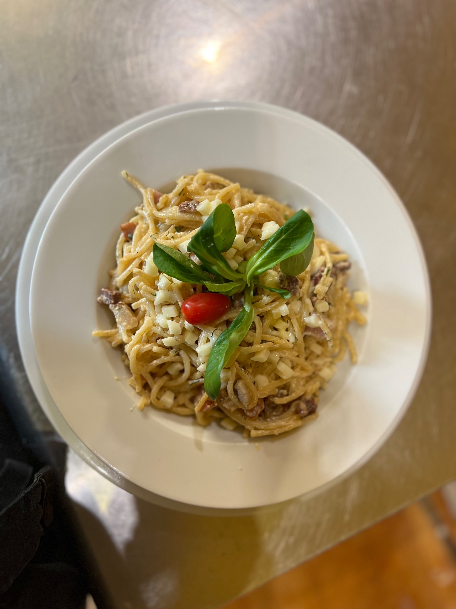 Spaghetti carbonara na białym talerzu, udekorowane listkami roszponki i połówką pomidora koktajlowego, widoczne kawałki boczku i jajka.