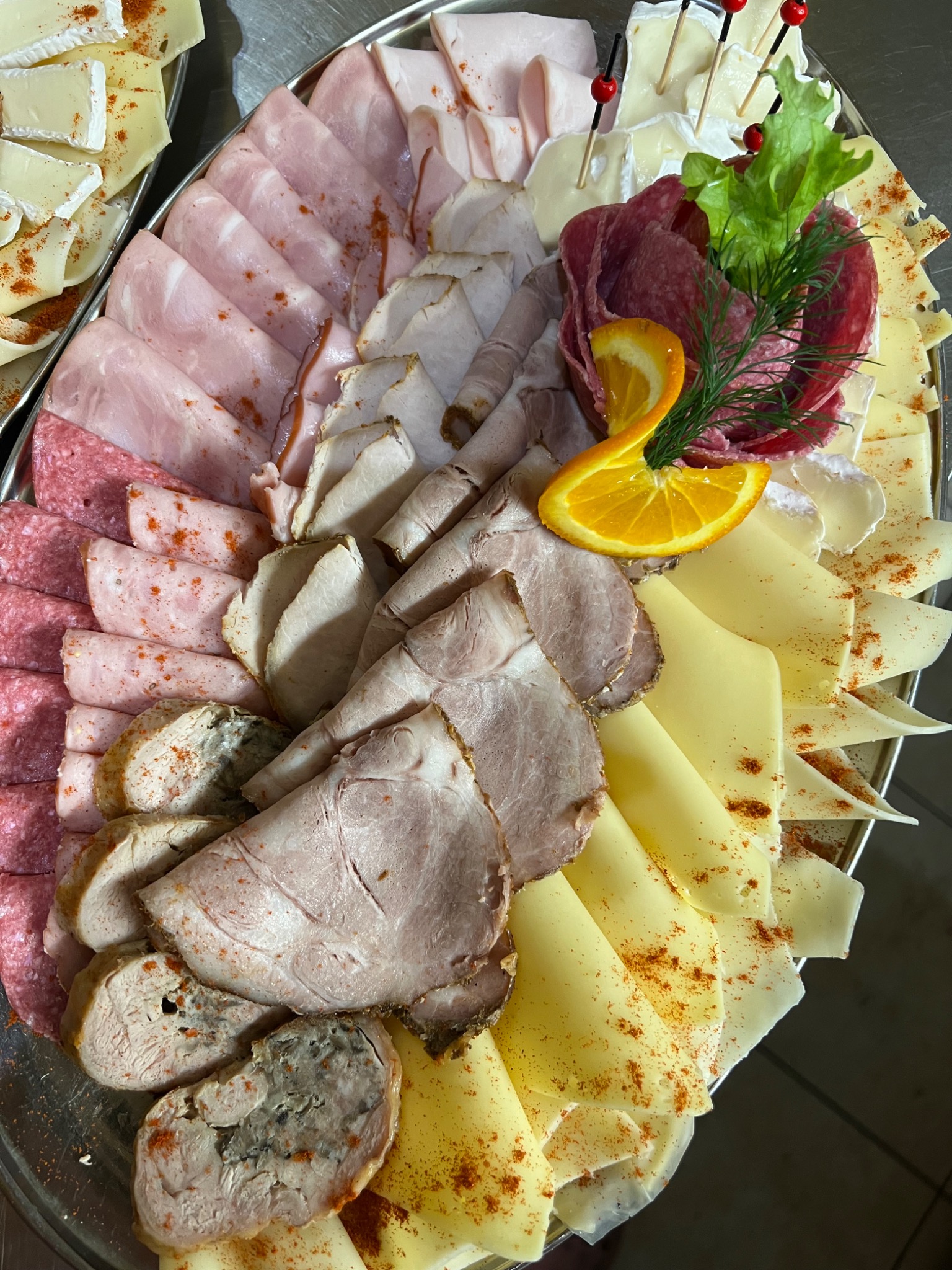 Elegancko ułożona na srebrnej tacy kompozycja wędlin i serów, udekorowana pomarańczą i zieleniną, posypana przyprawą.