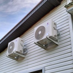 Przedsiębiorstwo usługowe Dawid Domagała - Dwa białe klimatyzatory marki Gree zamontowane na ścianie budynku z jasną elewacją, widoczne przewody i uchwyty montażowe, okno z odbijającym się otoczeniem w tle, pochmurne niebo.