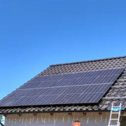 Panele fotowoltaiczne na dachu domu jednorodzinnego, drabina oparta o ścianę, niebieskie niebo w tle.