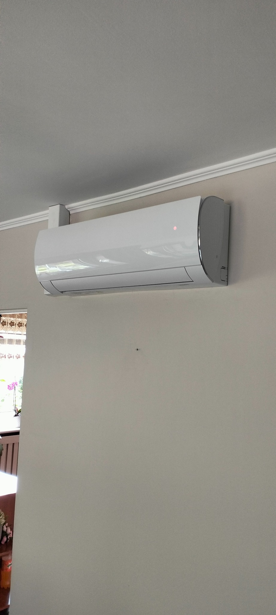 Biały klimatyzator marki Gree zamontowany na ścianie w jasnym pomieszczeniu, widoczna dioda sygnalizacyjna i fragment okna z kwiatami w tle.