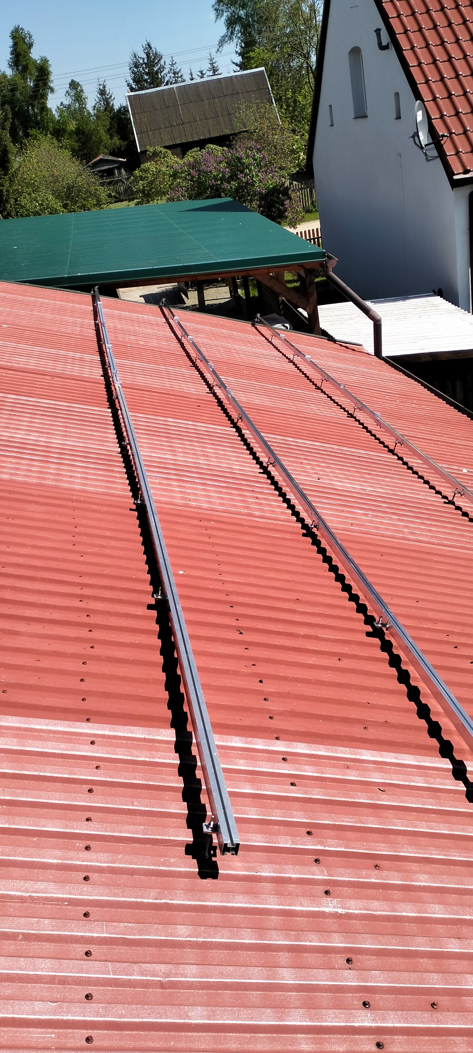 Montaż szyn pod panele fotowoltaiczne na czerwonym, trapezowym dachu z widocznymi elementami mocującymi i fragmentami budynków w tle.