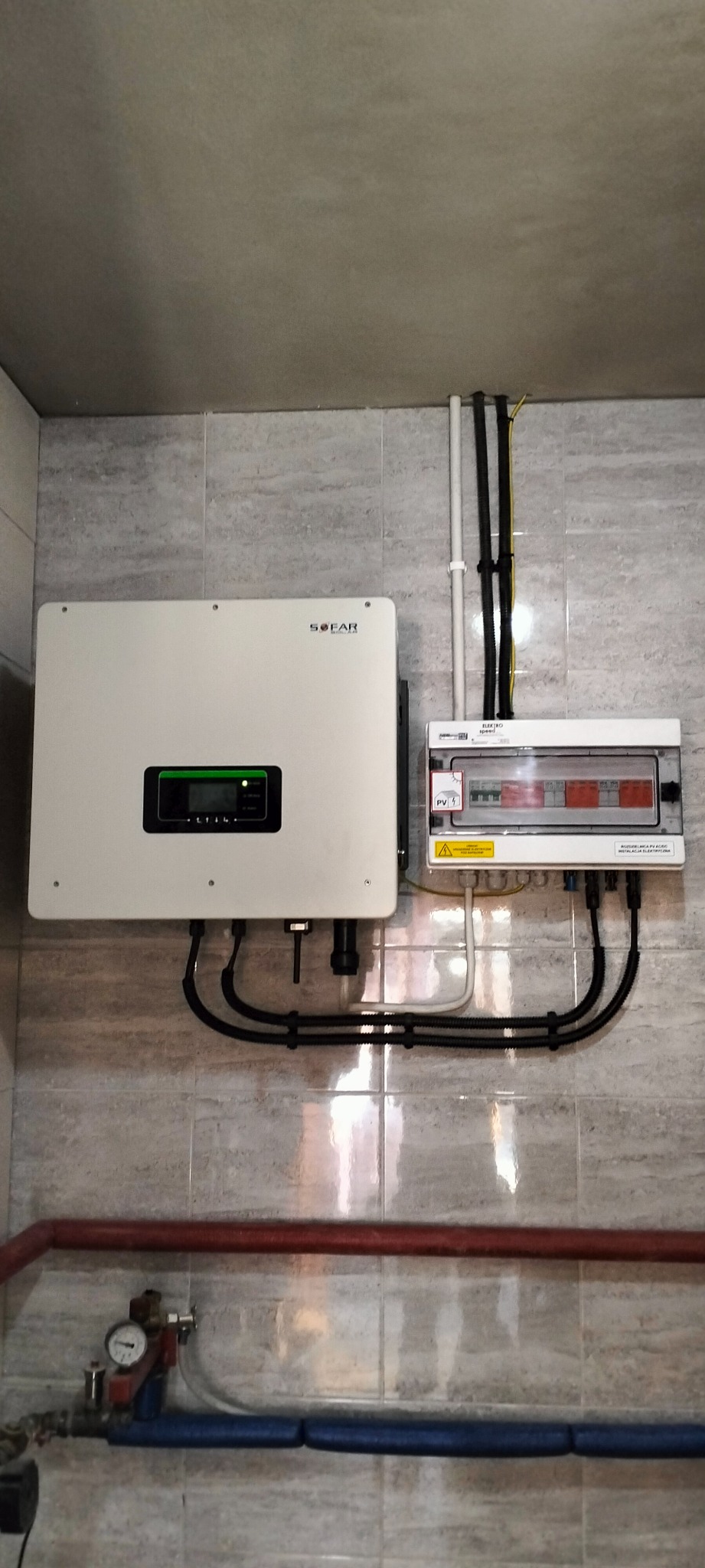 Zamontowany inwerter fotowoltaiczny marki Sofar Solar oraz rozdzielnica elektryczna z zabezpieczeniami na ścianie wyłożonej płytkami imitującymi kamień, widoczne rury instalacji centralnego...
