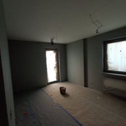 SAFE HANDS Valerii Teliatnikov - Pokój w trakcie przygotowań do malowania: szare ściany, zabezpieczona folią podłoga, zakryte okno, wiadro z farbą przy drzwiach, wystające przewody z sufitu.