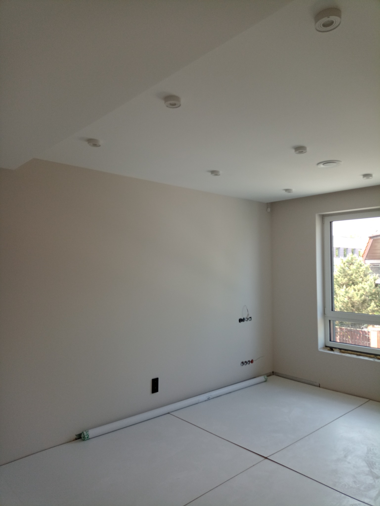 Pokój w trakcie prac wykończeniowych, widoczne przygotowane podłoże pod panele, świeżo pomalowane ściany w odcieniach beżu, sufit z zamontowanymi punktami oświetleniowymi i wystające kable...